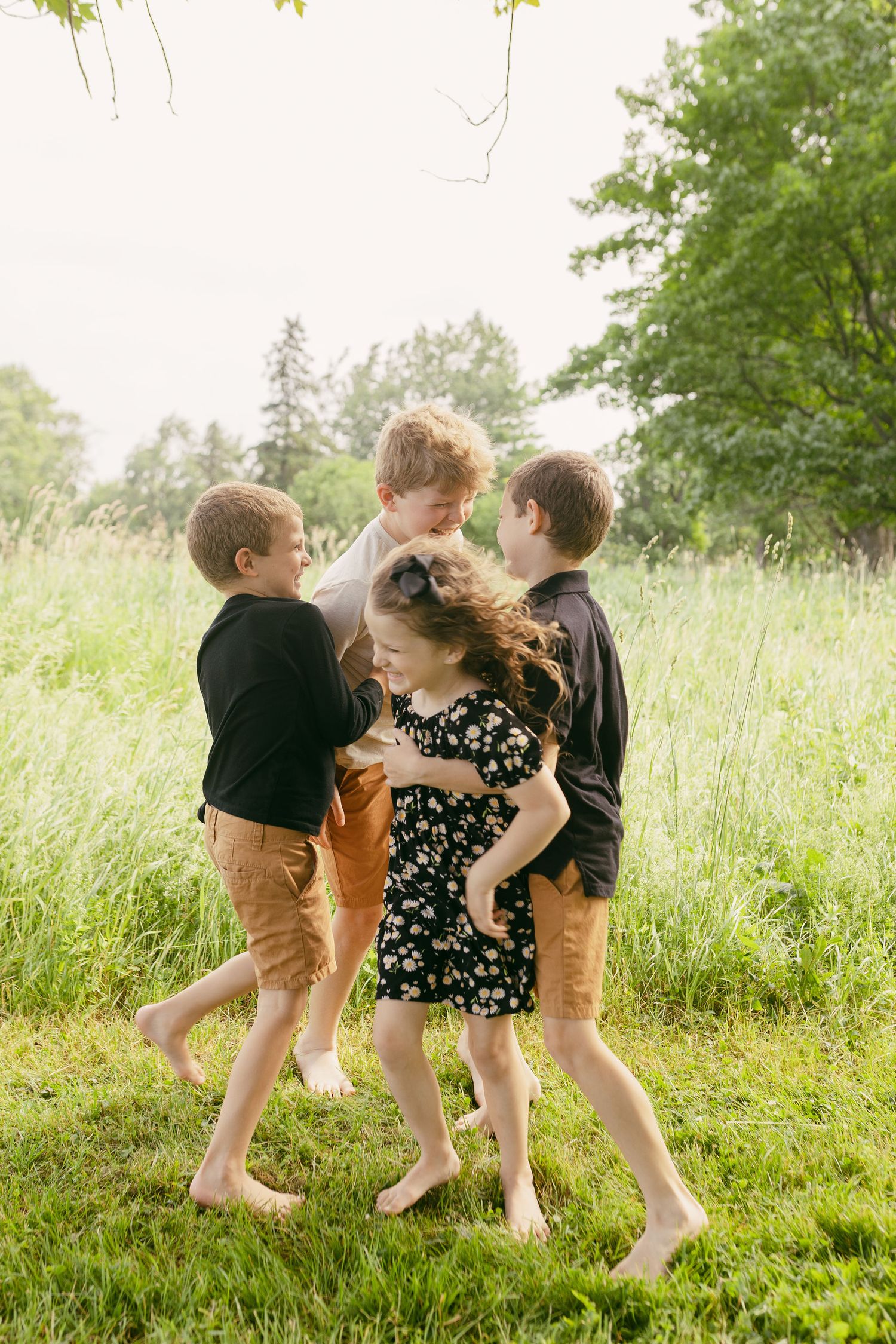 Four barefoot children playing and hugging together on a green grass field surrounded by trees on a sunny day.