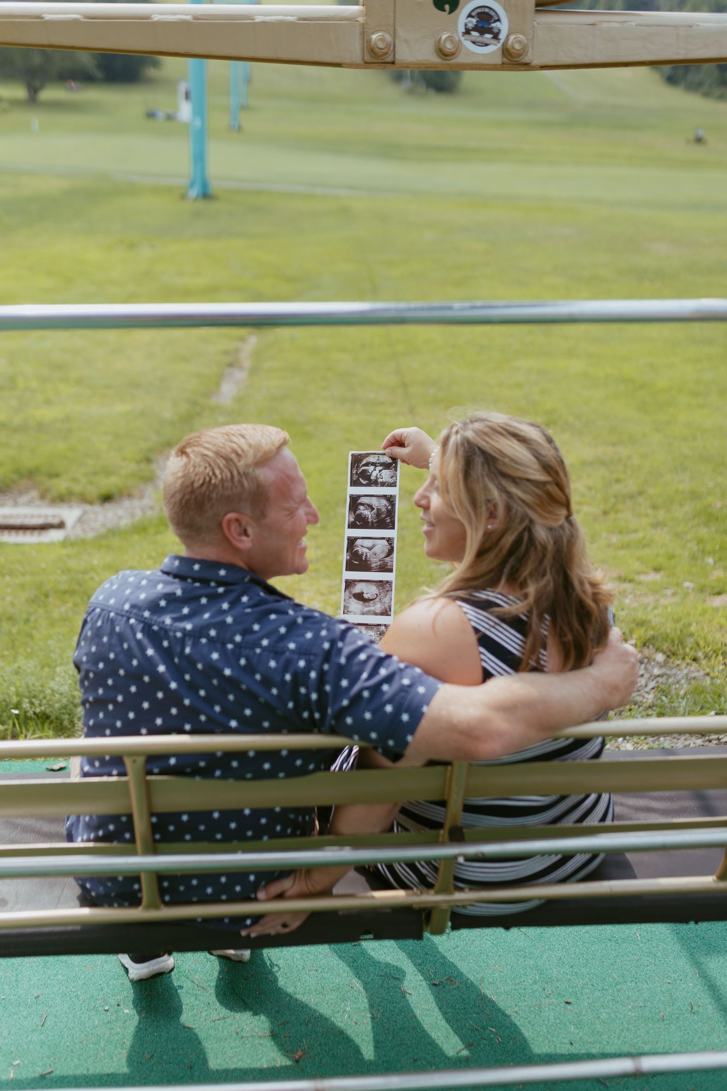 Couple sitting on a bench holding ultrasound photos, announcing pregnancy outdoors on a sunny day.