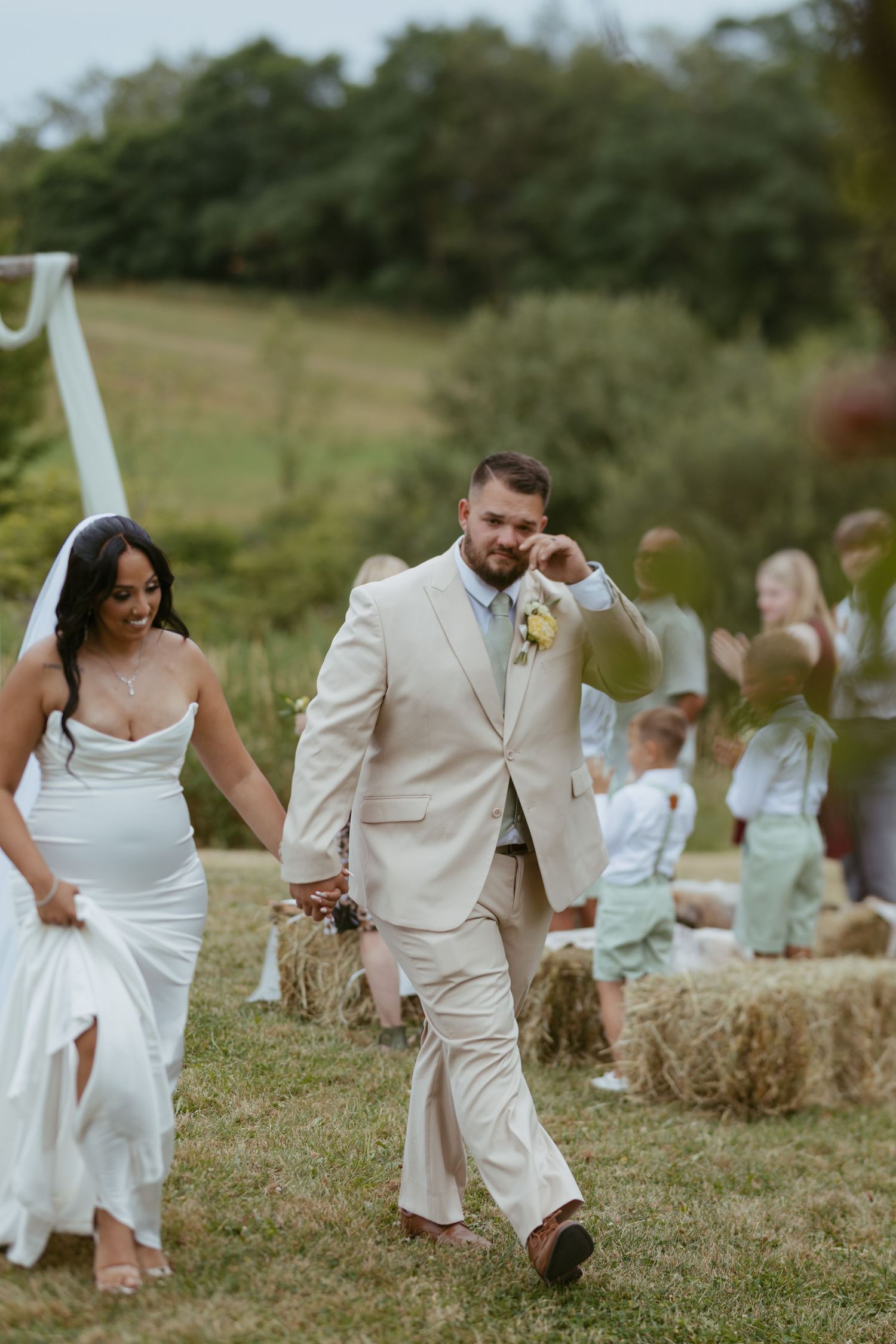 Bride and groom walk hand-in-hand down outdoor aisle lined with hay bales and guests at rustic wedding.