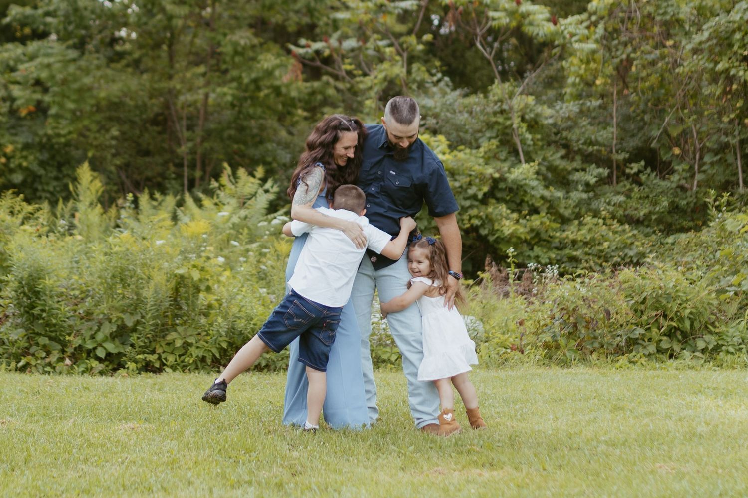 Family of four hugging and laughing together outdoors on green grass with lush garden background.