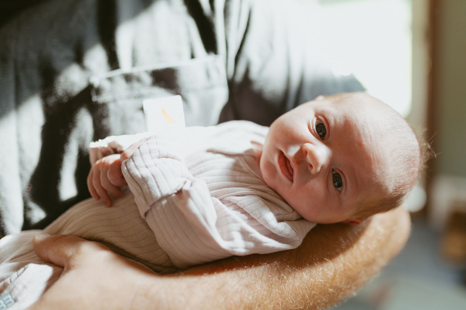 Newborn baby in white onesie cradled gently in adult hands, looking up with bright eyes in soft natural light.