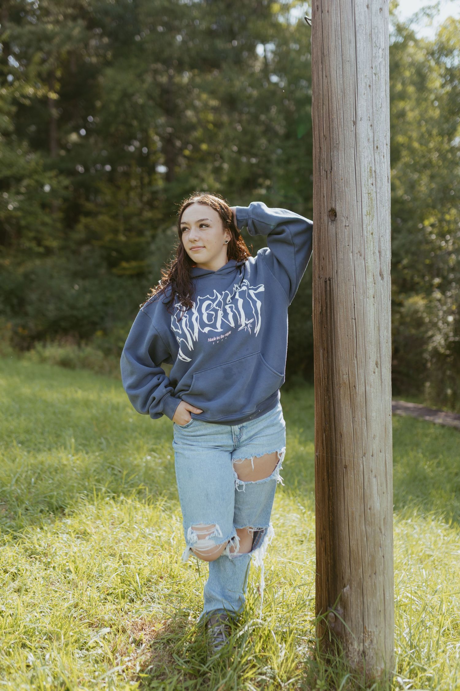 Young woman in ripped jeans and navy graphic sweatshirt leaning against wooden post in grassy outdoor setting.