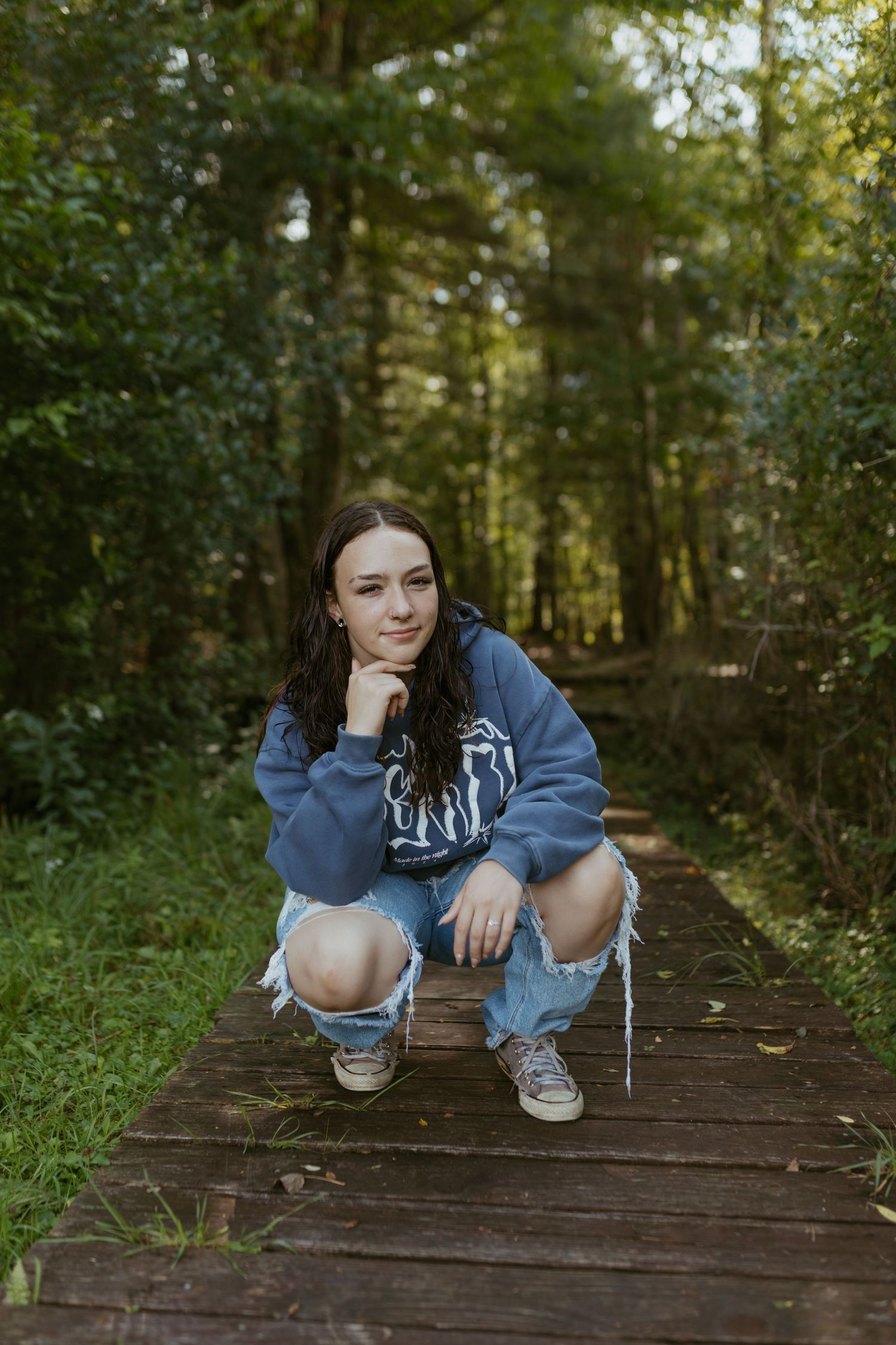 Young woman in blue hoodie and ripped jeans crouching on wooden path in lush green forest setting.