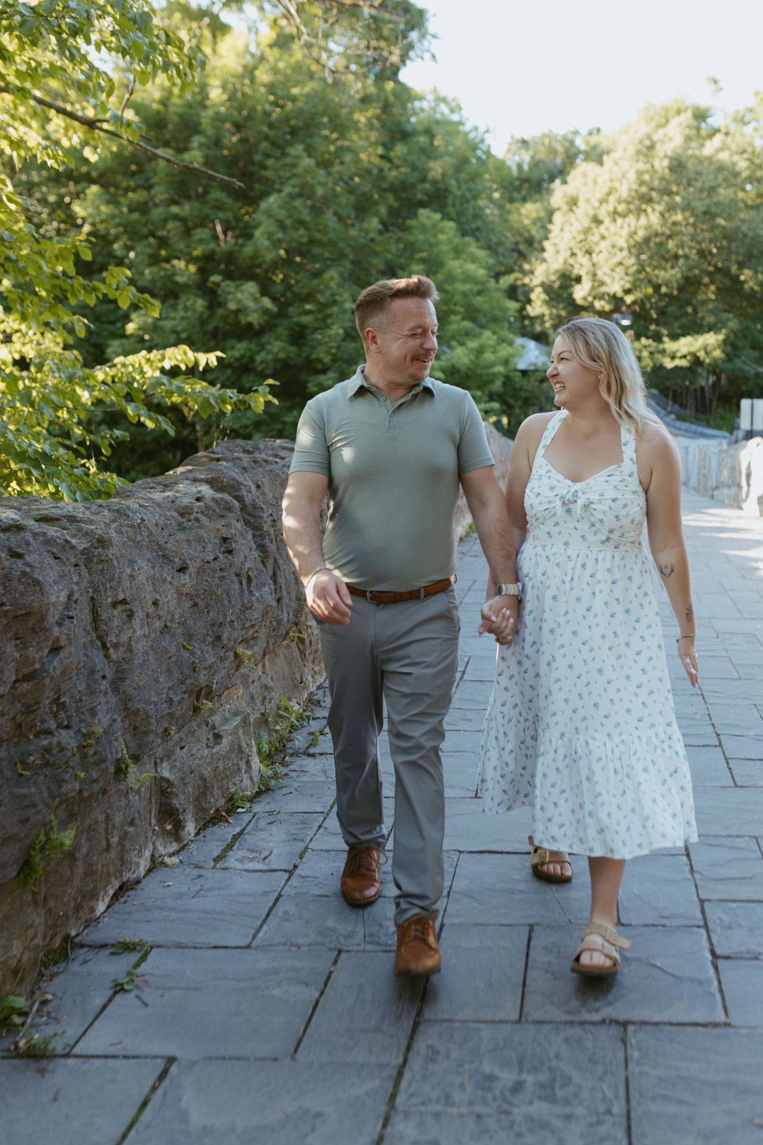 Happy couple holding hands walking on stone path surrounded by lush green trees.