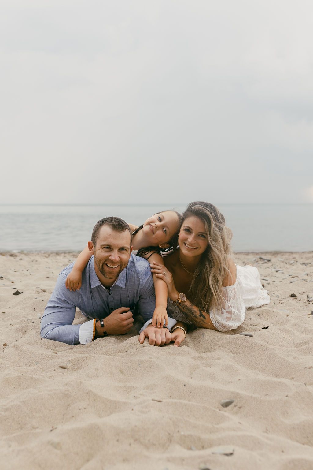 Family of three smiling and lying on sandy beach with ocean in background.