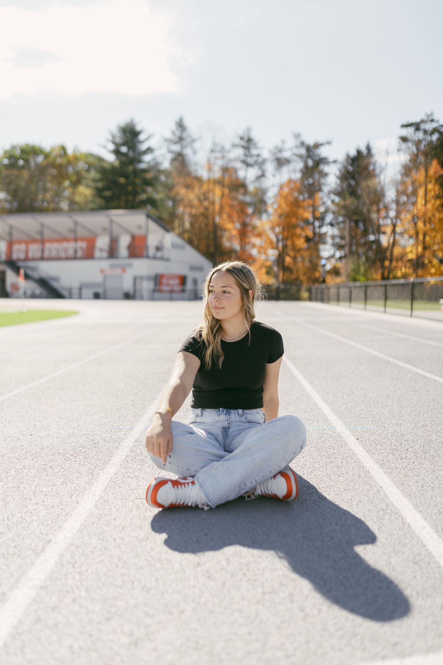 Young woman in black tee and jeans sits cross-legged on a running track with autumn trees in background.