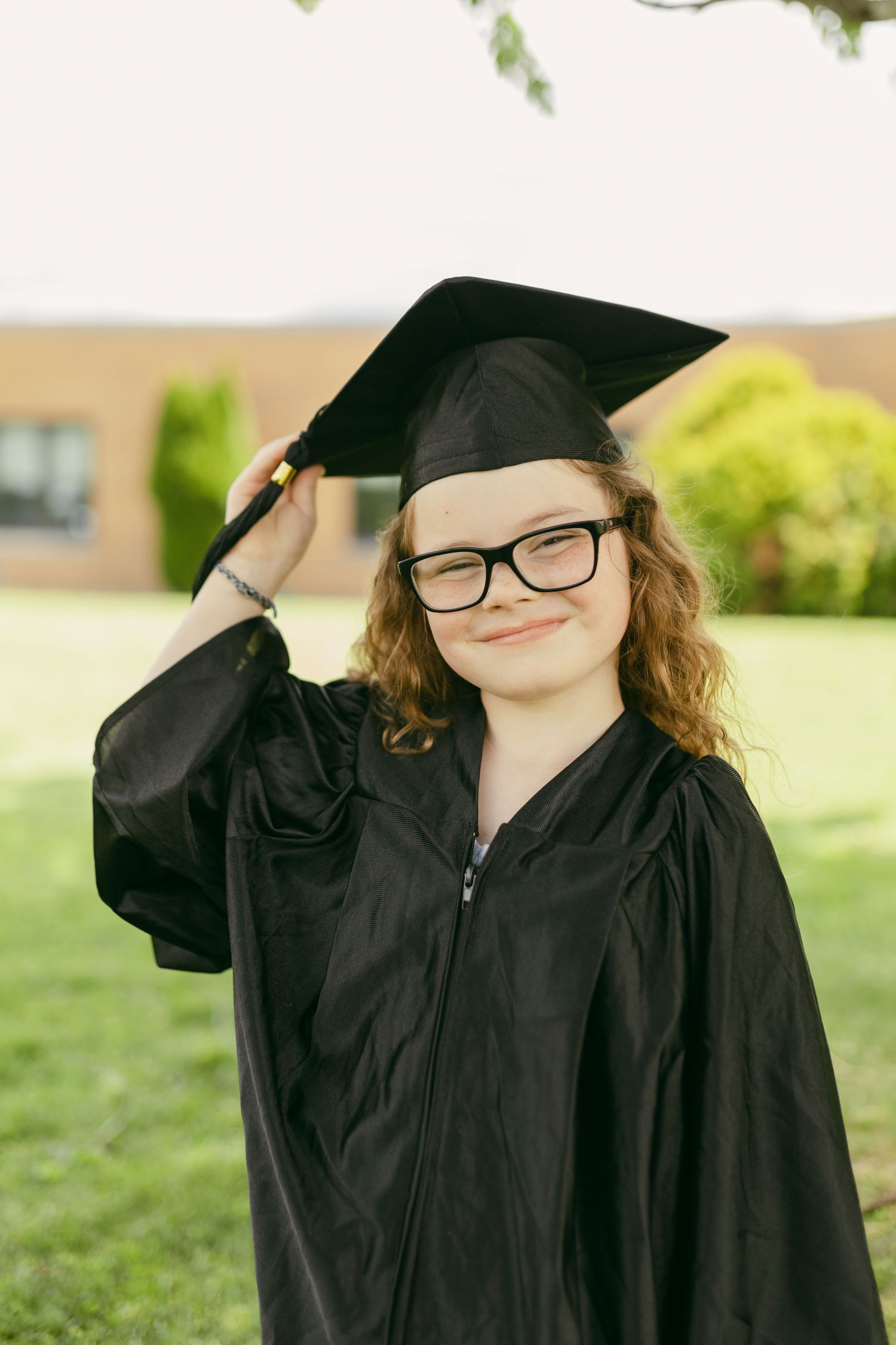 Young girl in black graduation cap and gown smiling outdoors, wearing glasses, celebrating academic achievement.