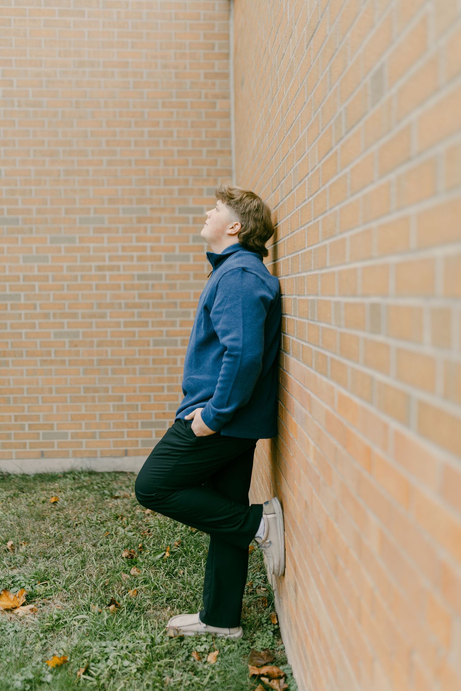 Young man in blue hoodie and black pants leaning against brick wall with one foot raised, looking upward outdoors.
