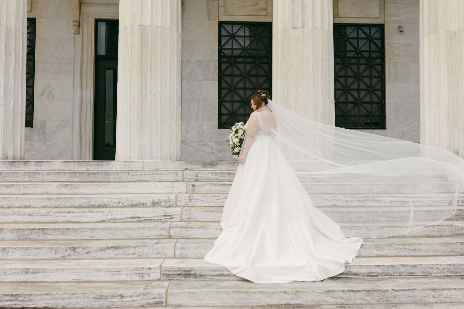 Bride in elegant white gown with long veil ascending marble steps of a grand columned building.