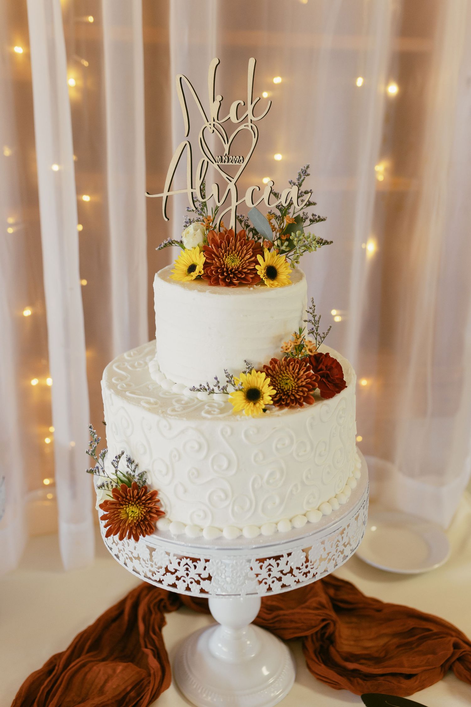 White wedding cake decorated with sunflowers and fall flowers on an elegant cake stand with fairy light backdrop.
