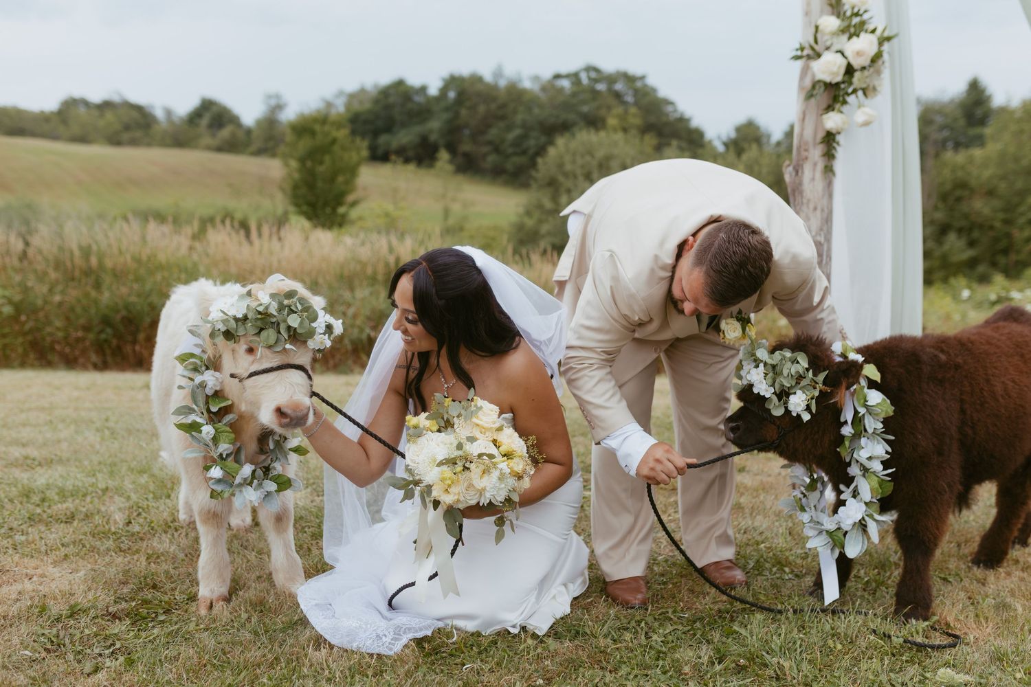 Bride and groom kneel with two flower crown-adorned mini horses at an outdoor farm wedding ceremony.