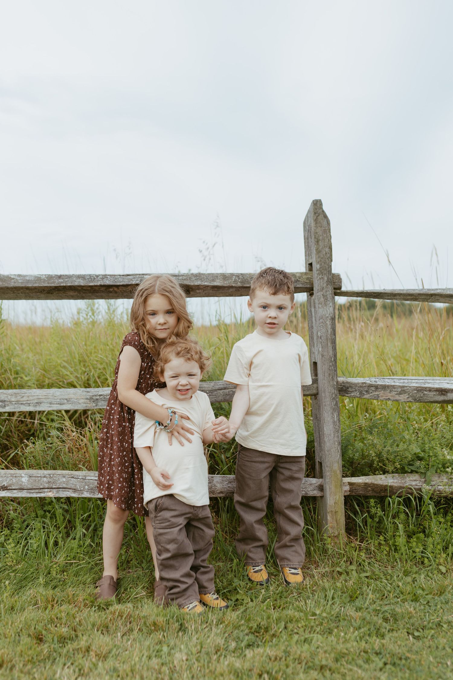 Three young children posing by a rustic wooden fence in a green field, wearing earth-toned outfits on a cloudy day.