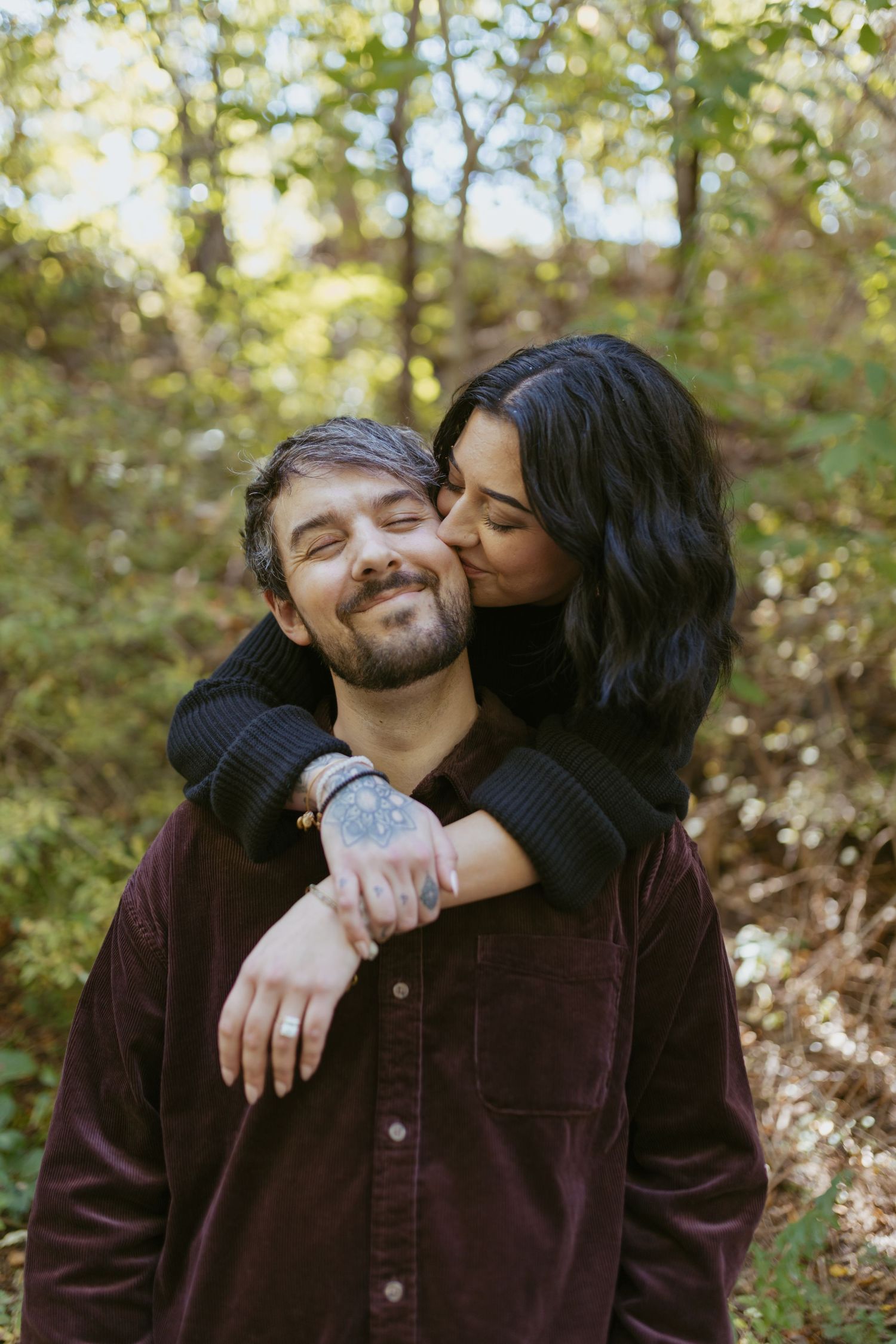 A woman with dark hair kisses a bearded man on the cheek outdoors in a lush green forest setting.