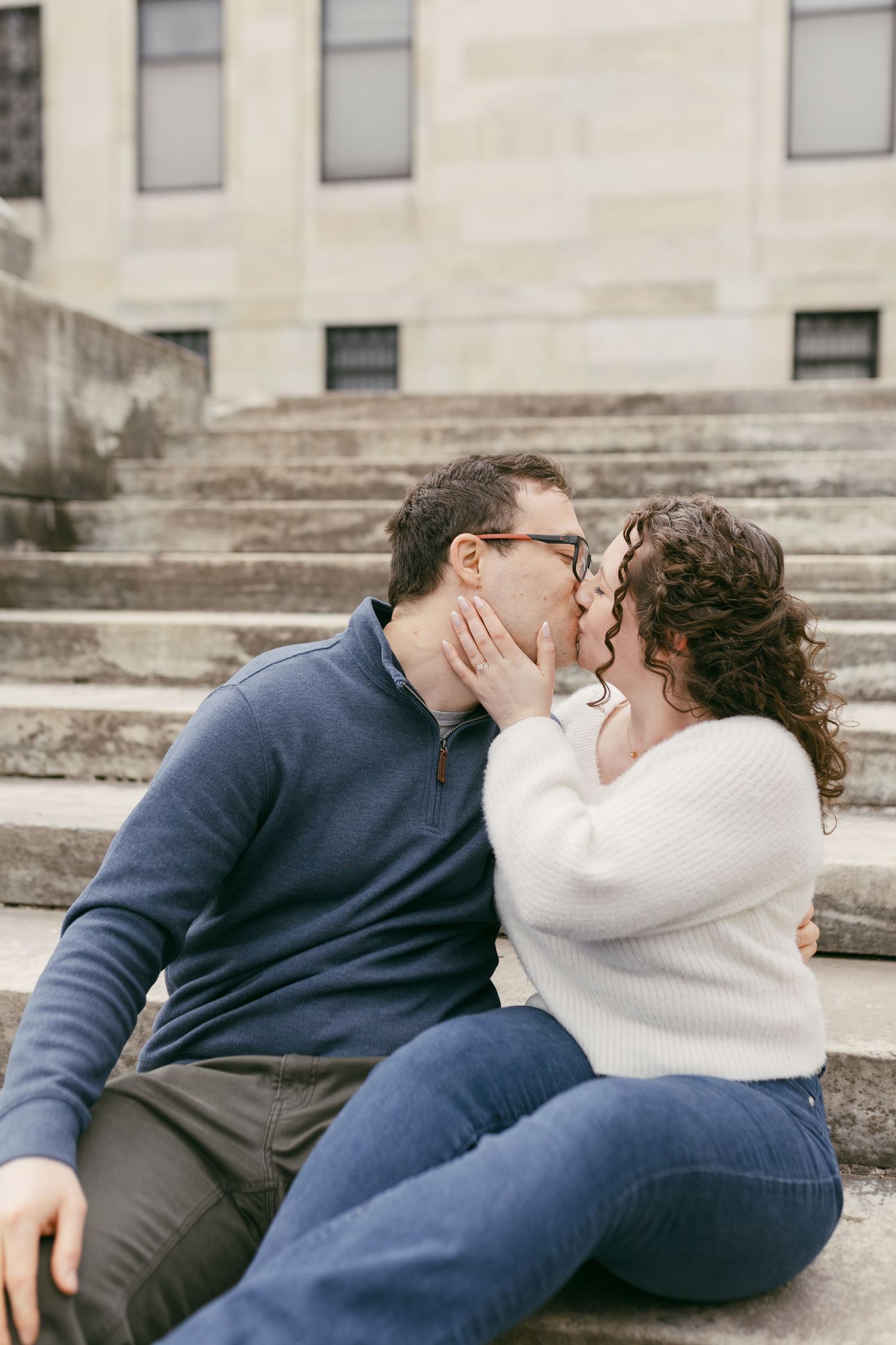 A couple shares a romantic kiss while sitting on stone steps outdoors, dressed in casual sweaters and jeans.