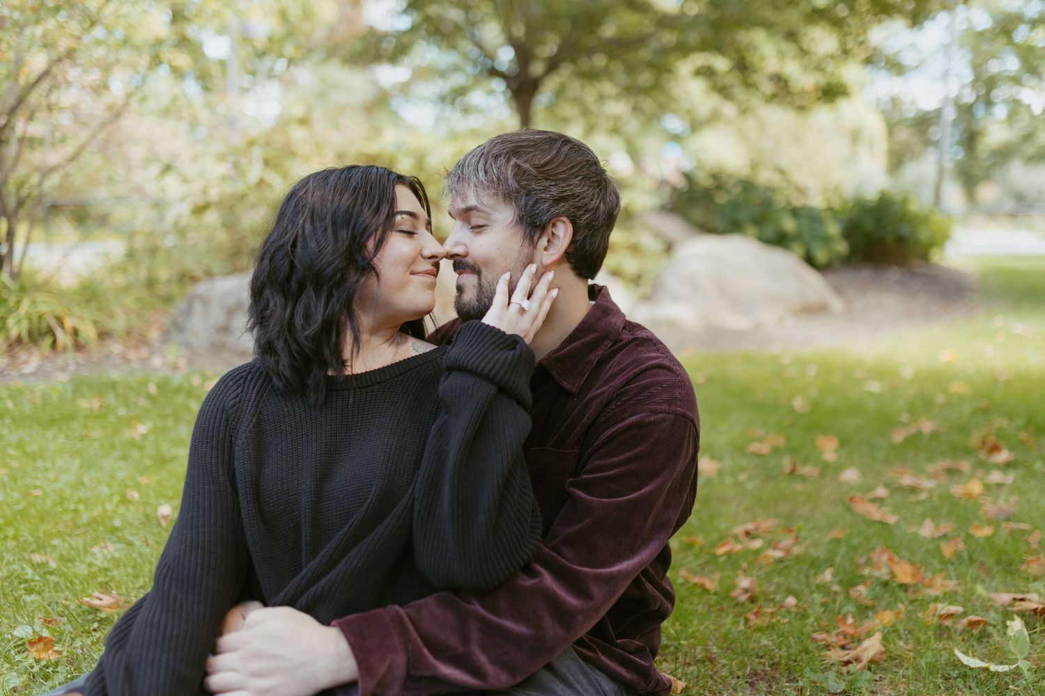 Couple sitting on grass in autumn park, woman touching man's face as they lean in for a kiss.