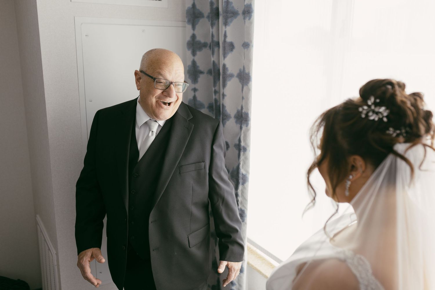An emotional older man in a gray suit reacts with joy upon seeing the bride during a first look moment.