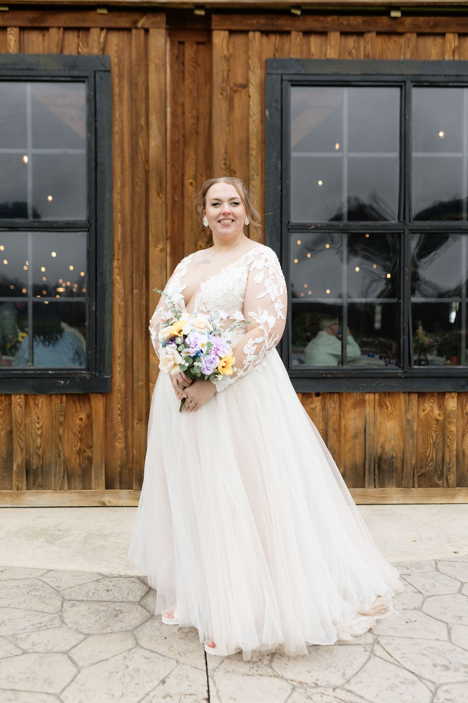 Bride in a long-sleeve lace tulle wedding gown holding a colorful bouquet, standing before a rustic wooden barn.