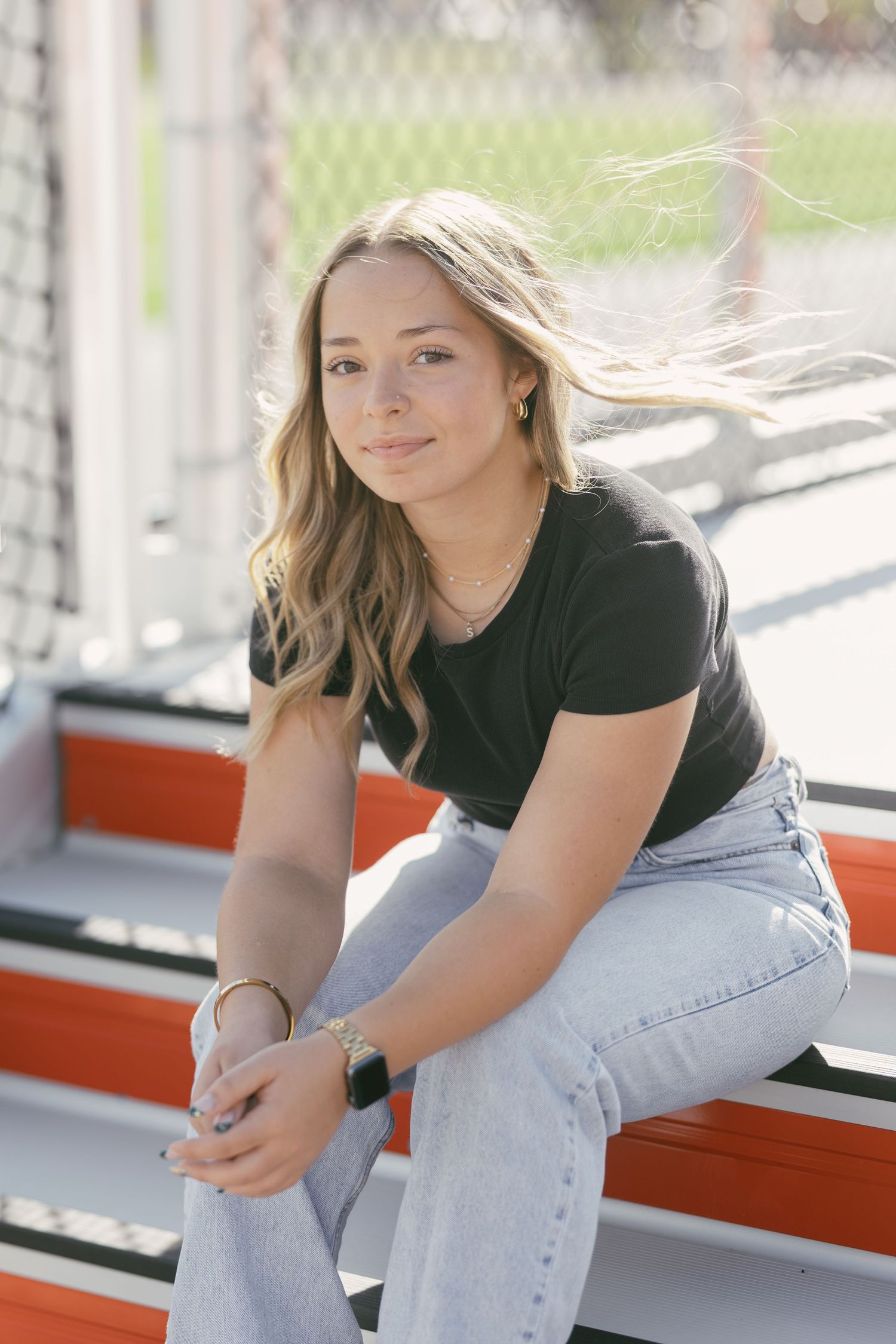 Young blonde woman in black tee and jeans sits on red bleachers outdoors, hair blowing in breeze.