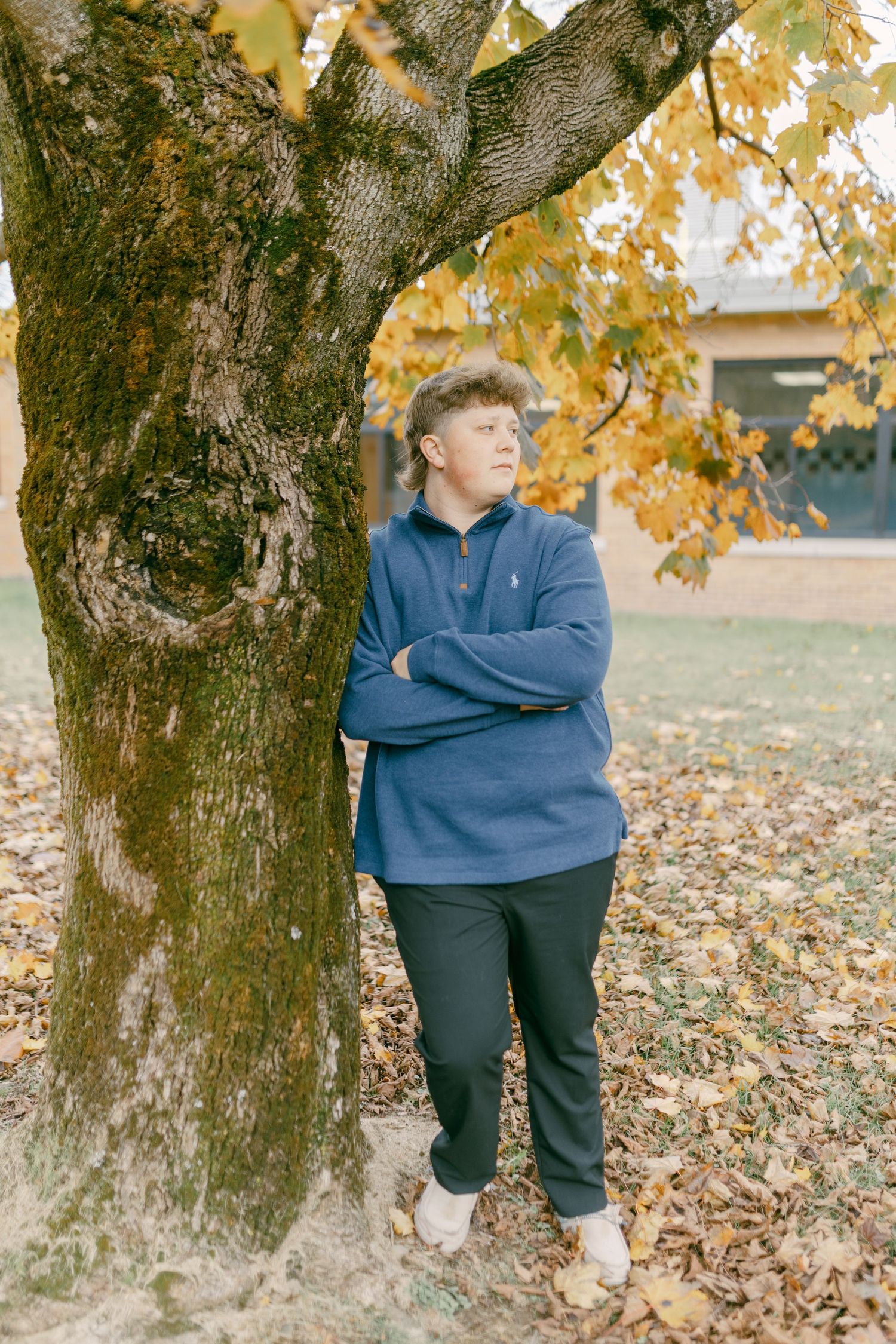Young man in blue pullover leaning against autumn tree with arms crossed, surrounded by fallen leaves.