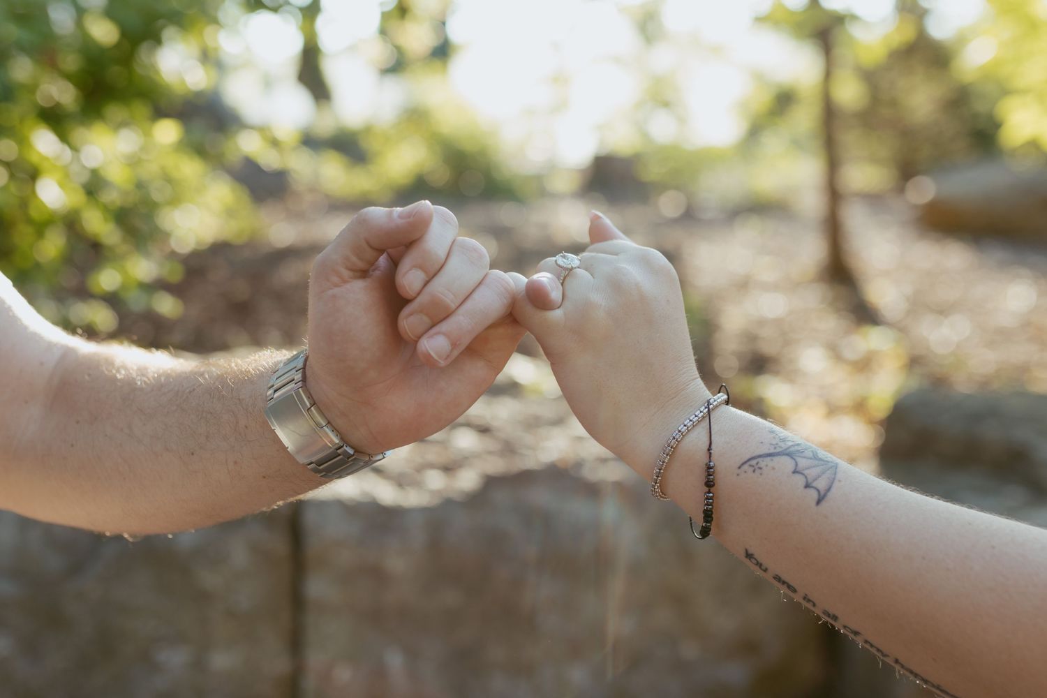Two hands linking pinky fingers outdoors, one wearing a silver bracelet and the other a delicate chain bracelet with a tattoo.