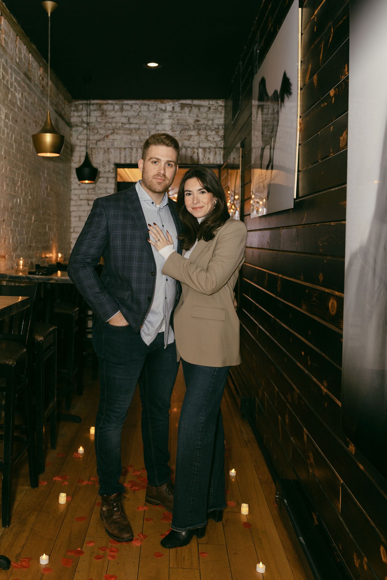 Couple posing romantically in candlelit restaurant with rose petals, brick walls, and wooden floors.