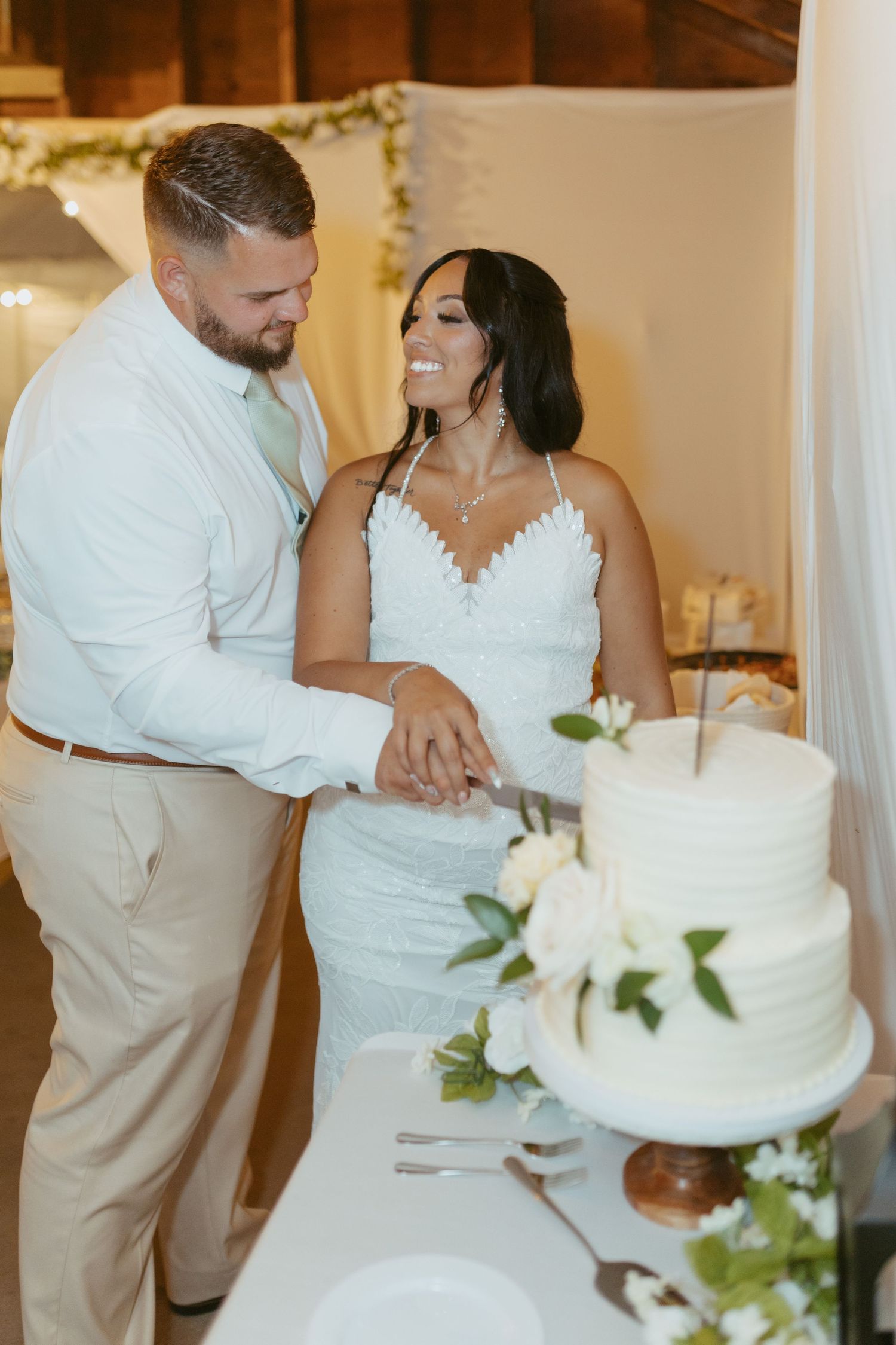 Bride and groom smiling while cutting their white two-tier wedding cake decorated with green leaves and white flowers.