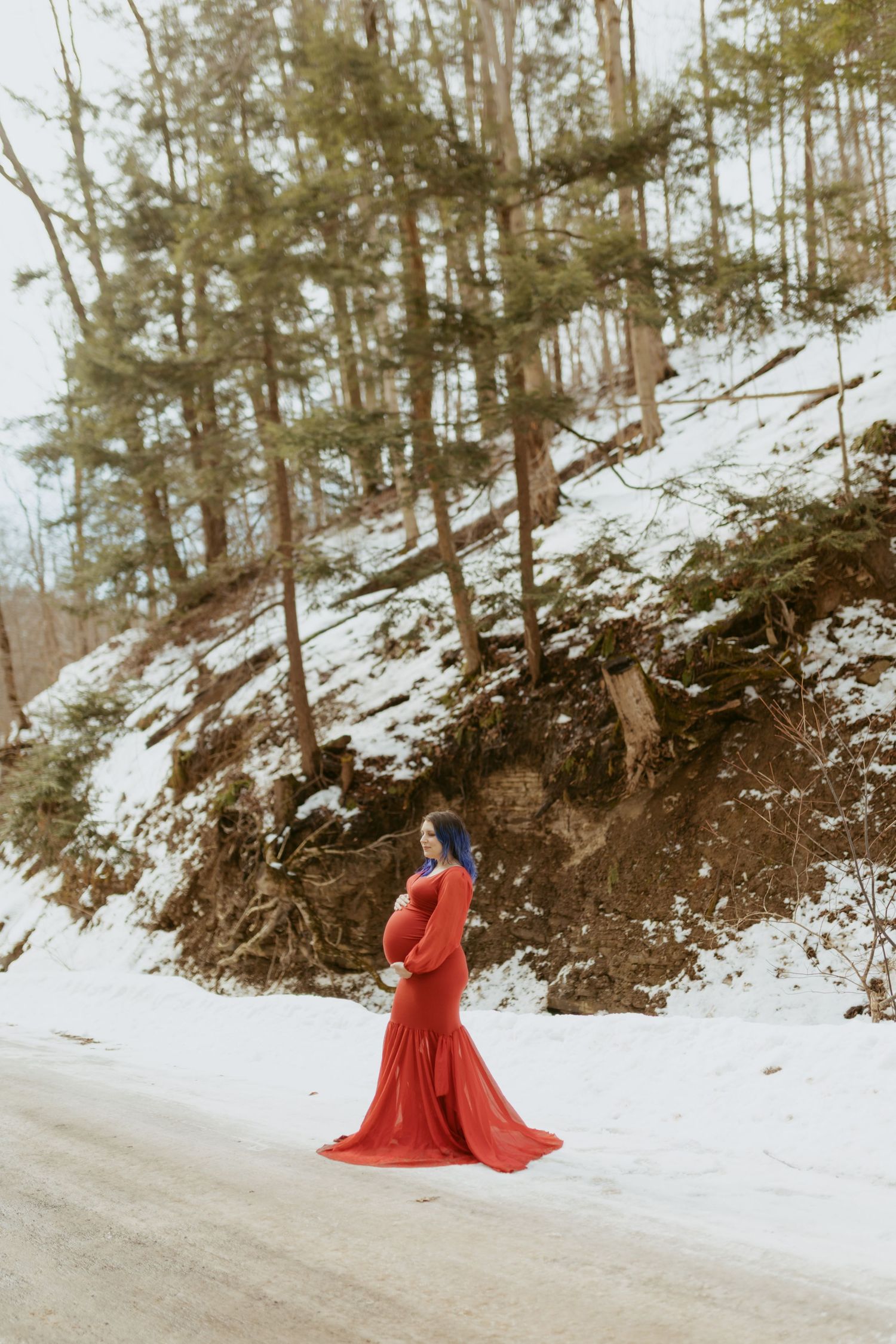 Pregnant woman in red gown standing on snowy road surrounded by pine trees in a winter maternity photo shoot.