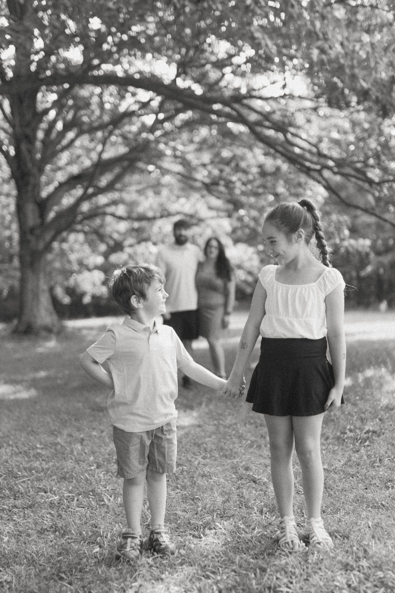 Black and white photo of two children holding hands in a park, with parents walking behind them among trees.