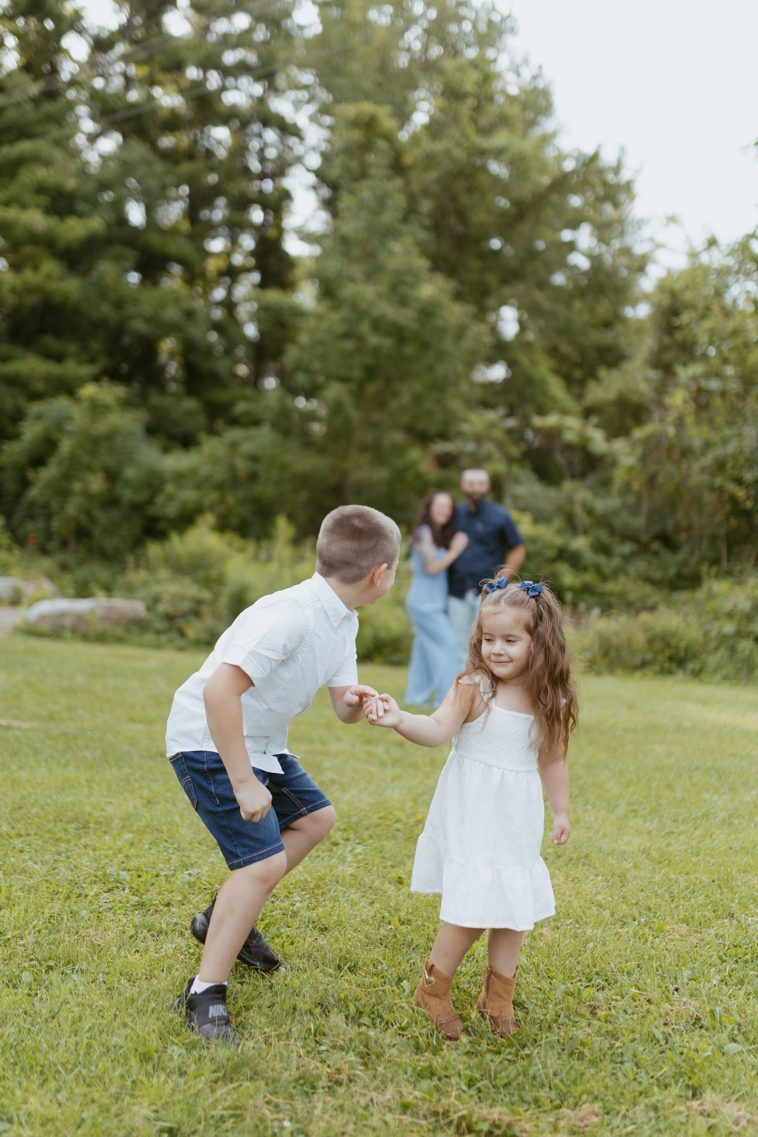 Two children hold hands and play in a green park while their parents watch lovingly in the background.