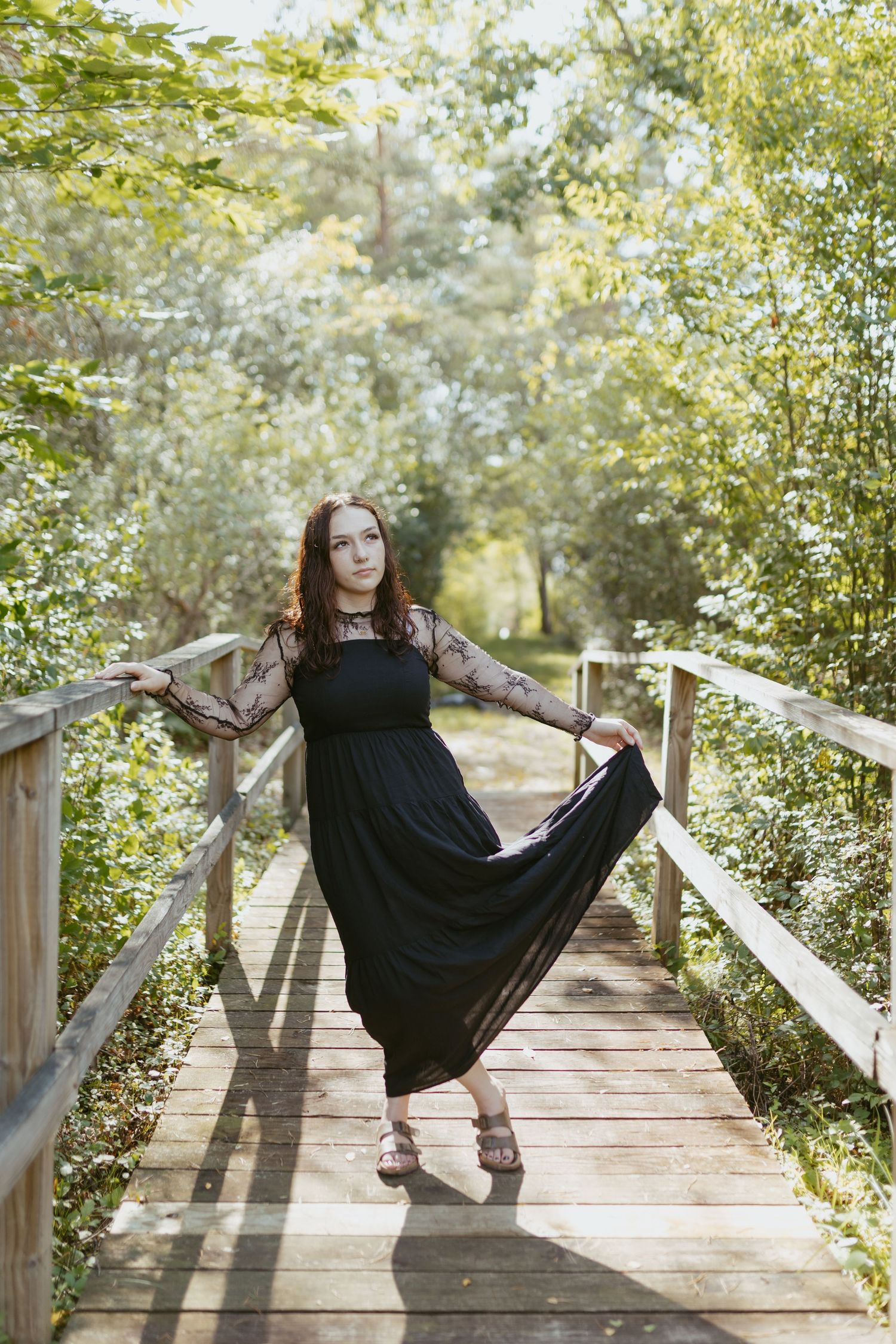 Young woman in elegant black lace dress posing on wooden bridge surrounded by lush green trees.