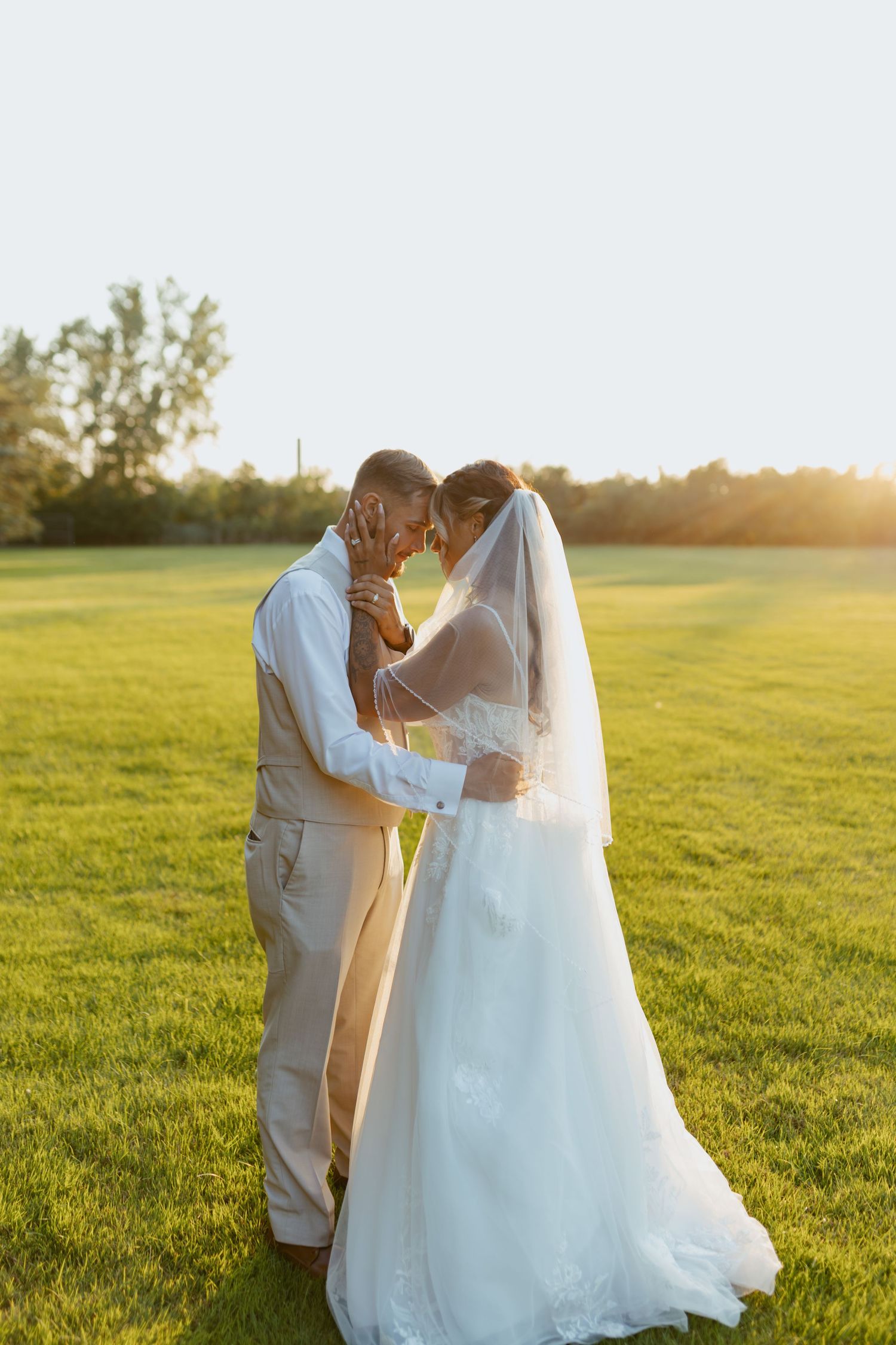 A bride and groom share a romantic moment at golden hour on a lush green field, foreheads touching tenderly.