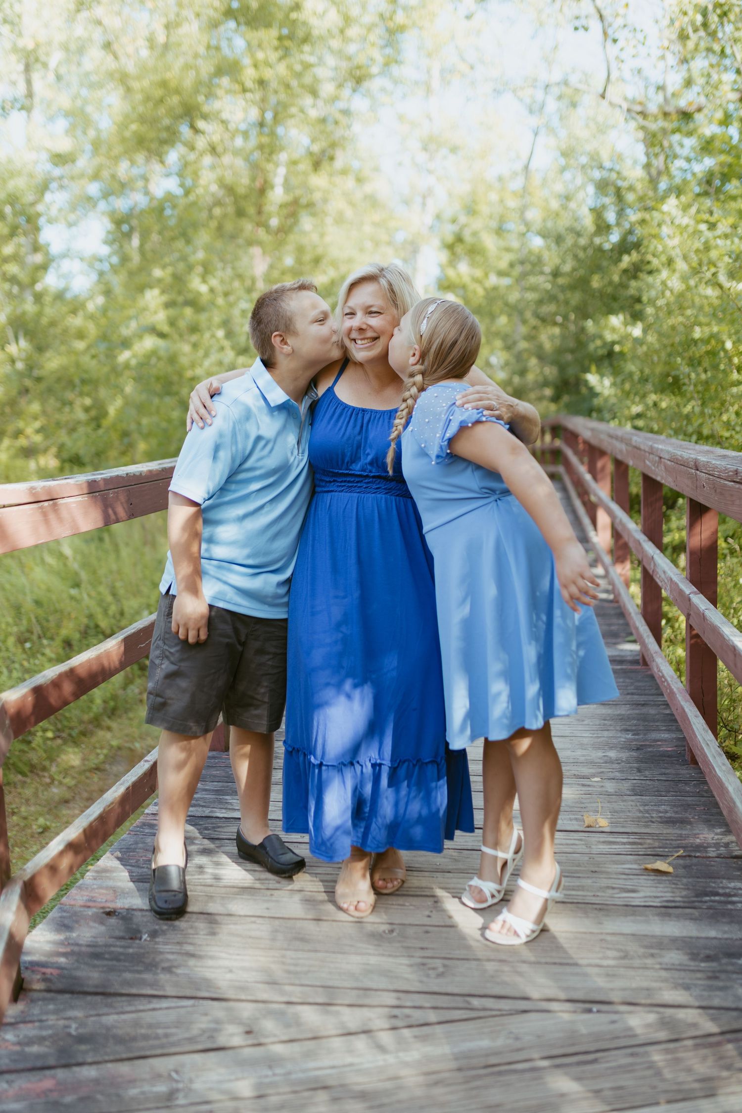A woman in a blue dress smiles on a wooden bridge as two children lean in to kiss her cheeks outdoors.
