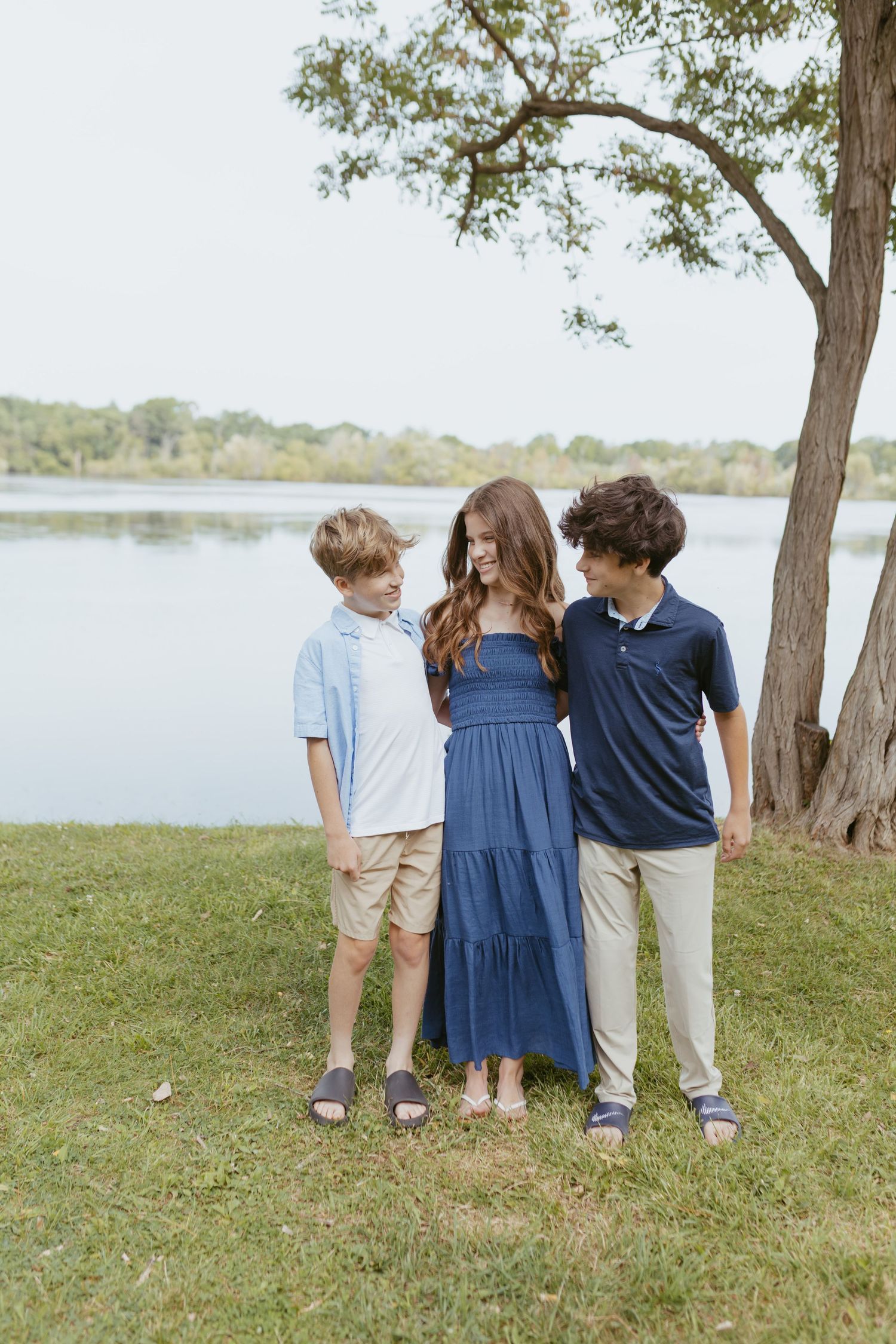 Three kids posing outdoors near a lake, wearing coordinated blue and khaki outfits on a grassy lawn by a tree.