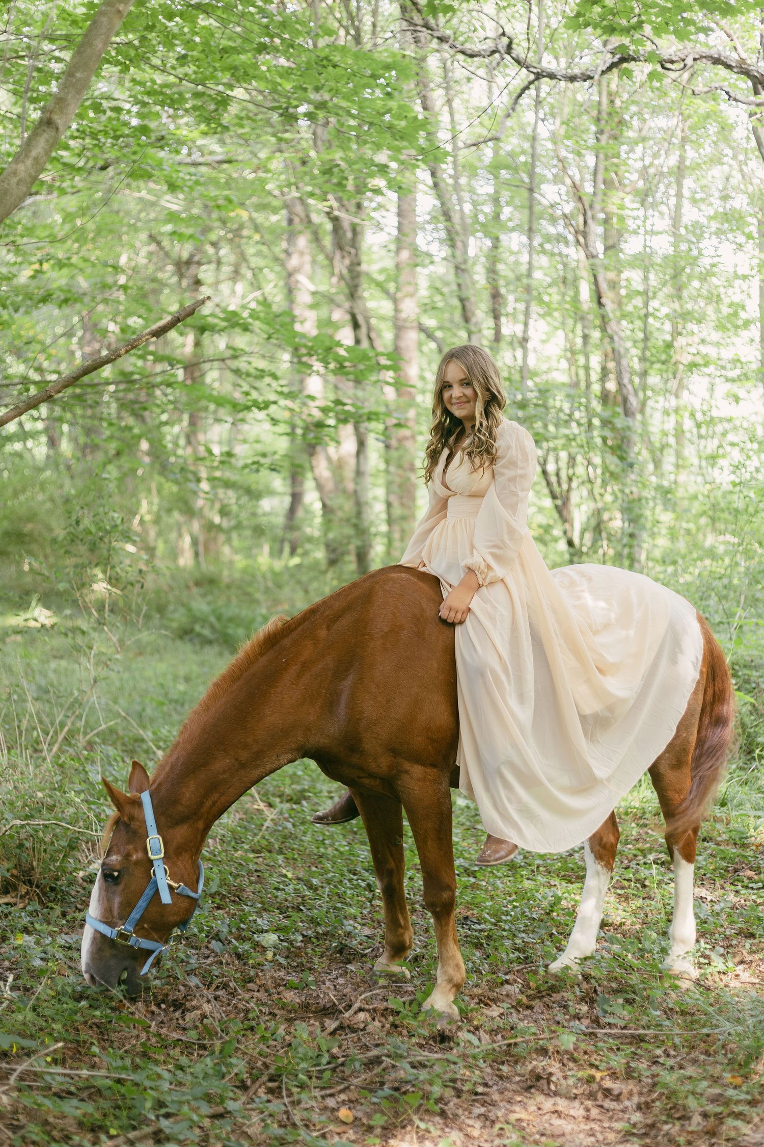 A brown horse and someone in a flowing cream dress stand together in a sunlit forest.