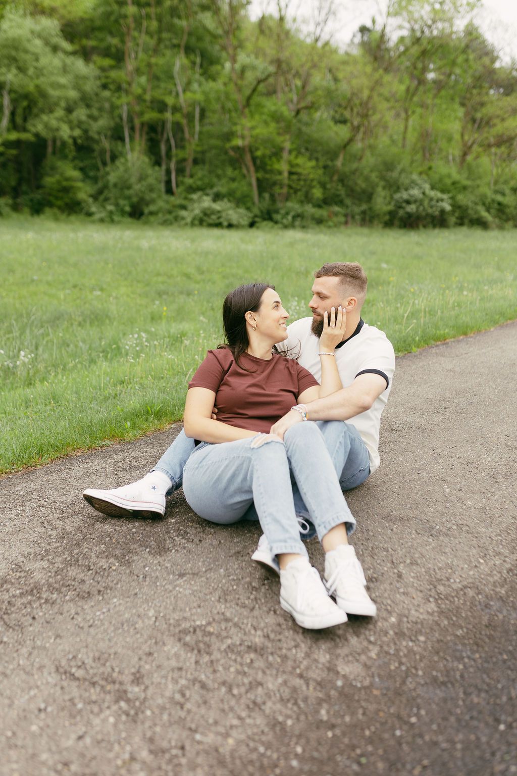 A couple sits intimately on a path surrounded by green grass and trees.