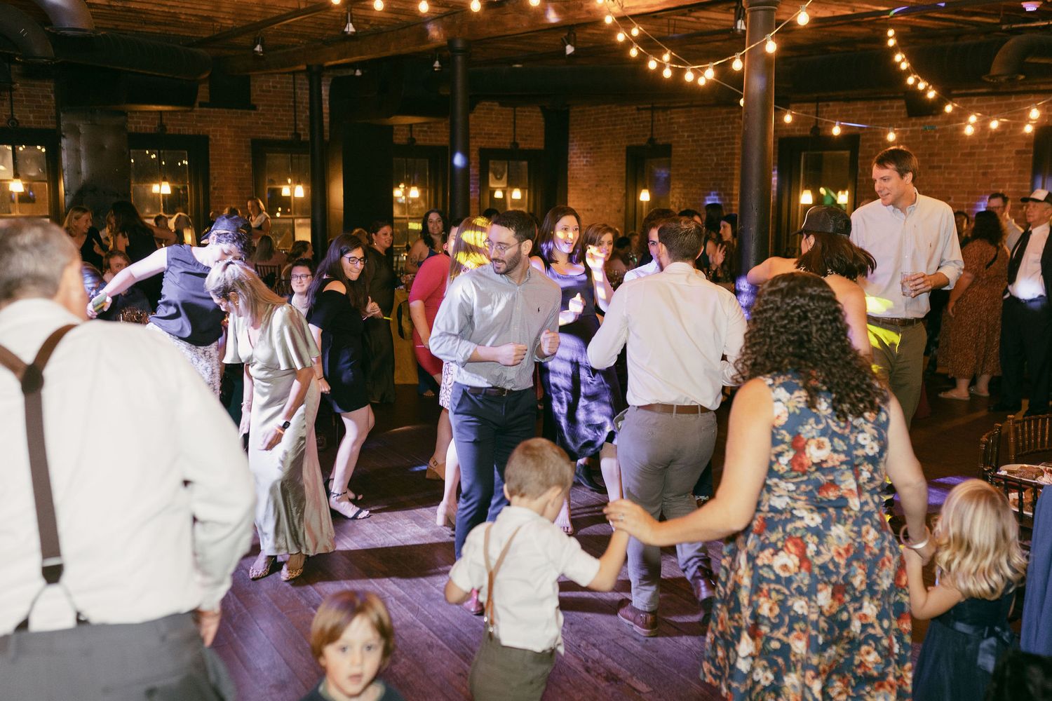 Wedding guests dancing and celebrating at reception under warm string lights.