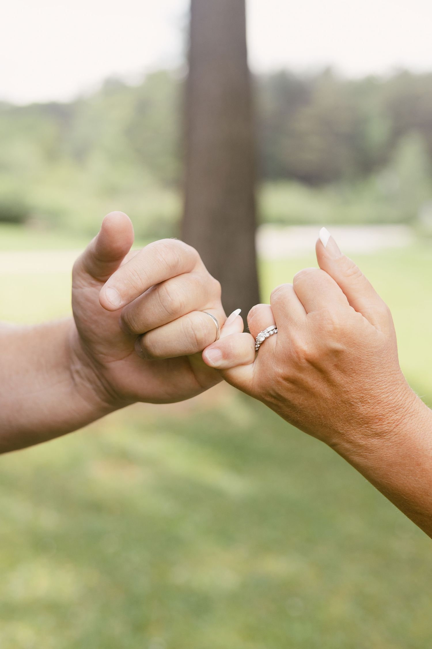 Close-up of hands with engagement ring linking pinky fingers together outdoors.