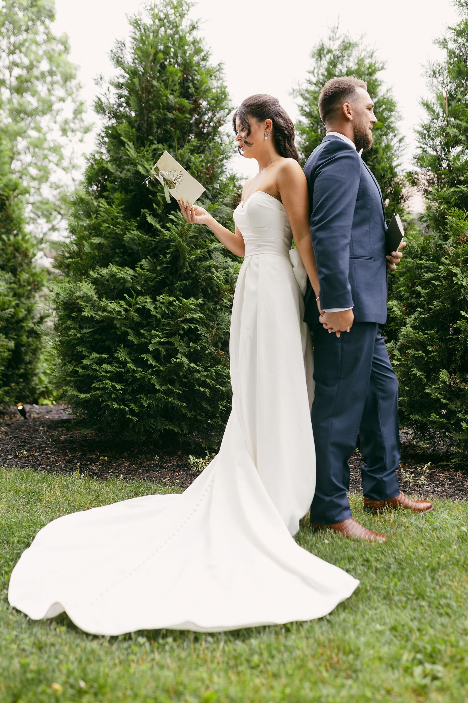 Elegant wedding dress train flows across green lawn with evergreen trees in background.