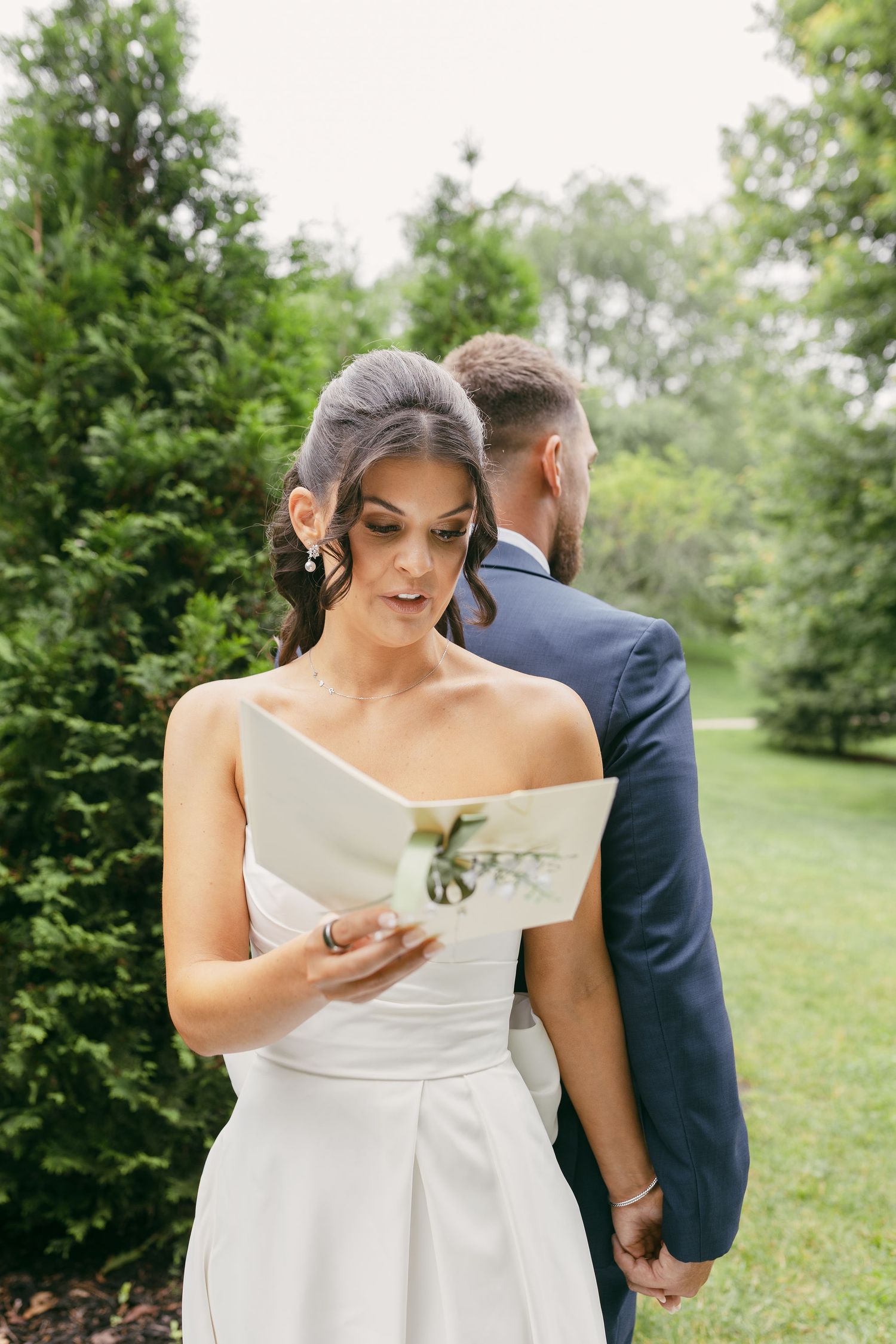 Person in white off-shoulder wedding dress reads from paper in outdoor garden setting.