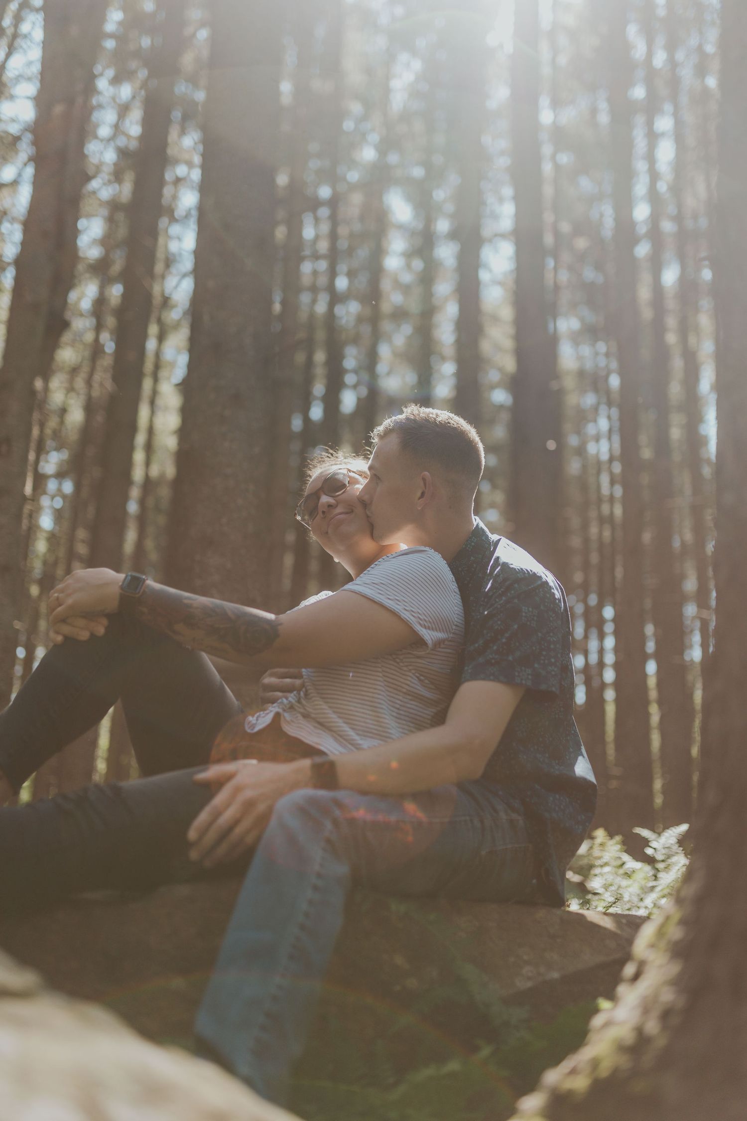 Couple sitting together on a rock in a sunlit forest, sharing a tender moment among tall pine trees.
