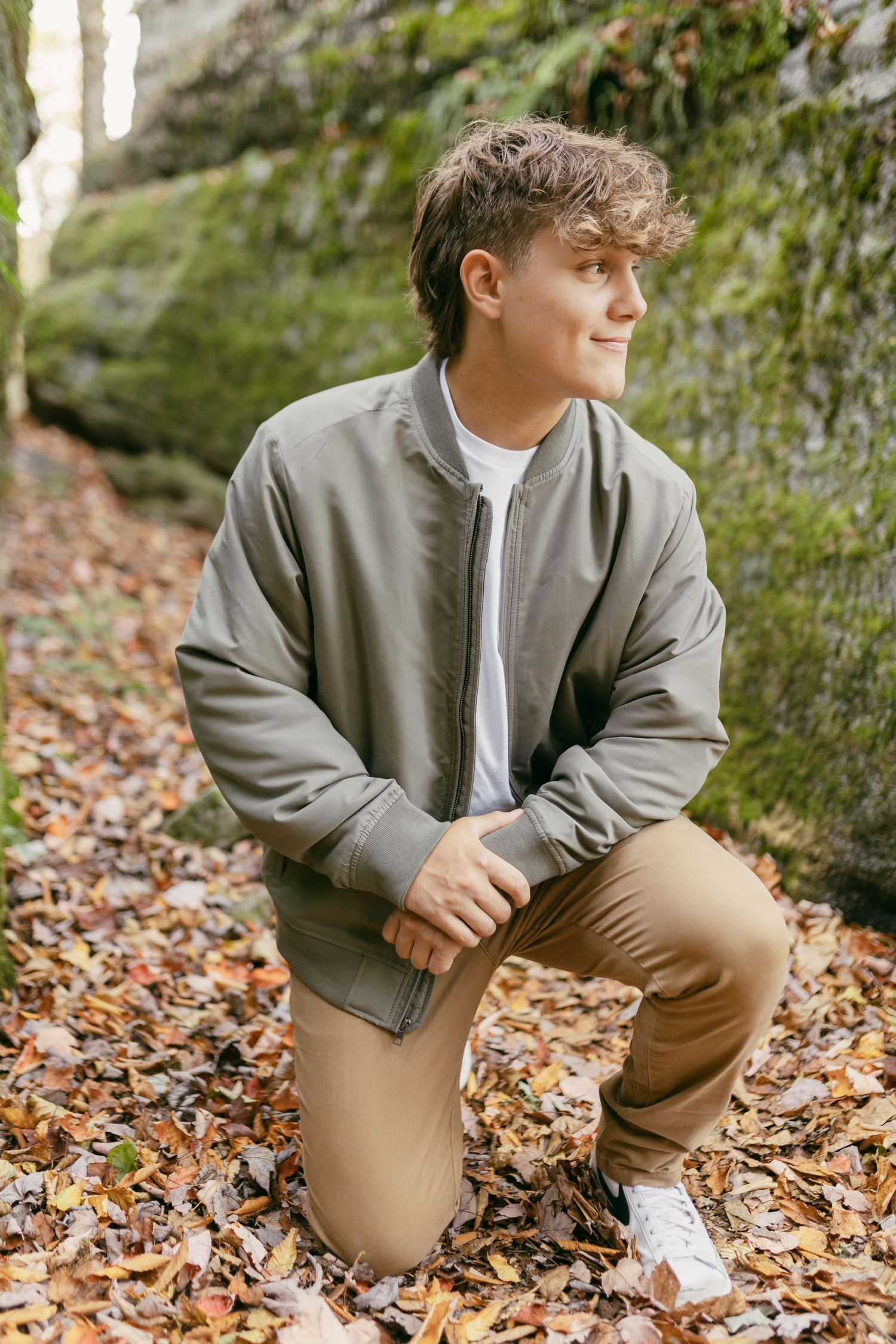 Young man kneeling on fall leaves near mossy rocks, wearing olive bomber jacket and tan pants, looking away.