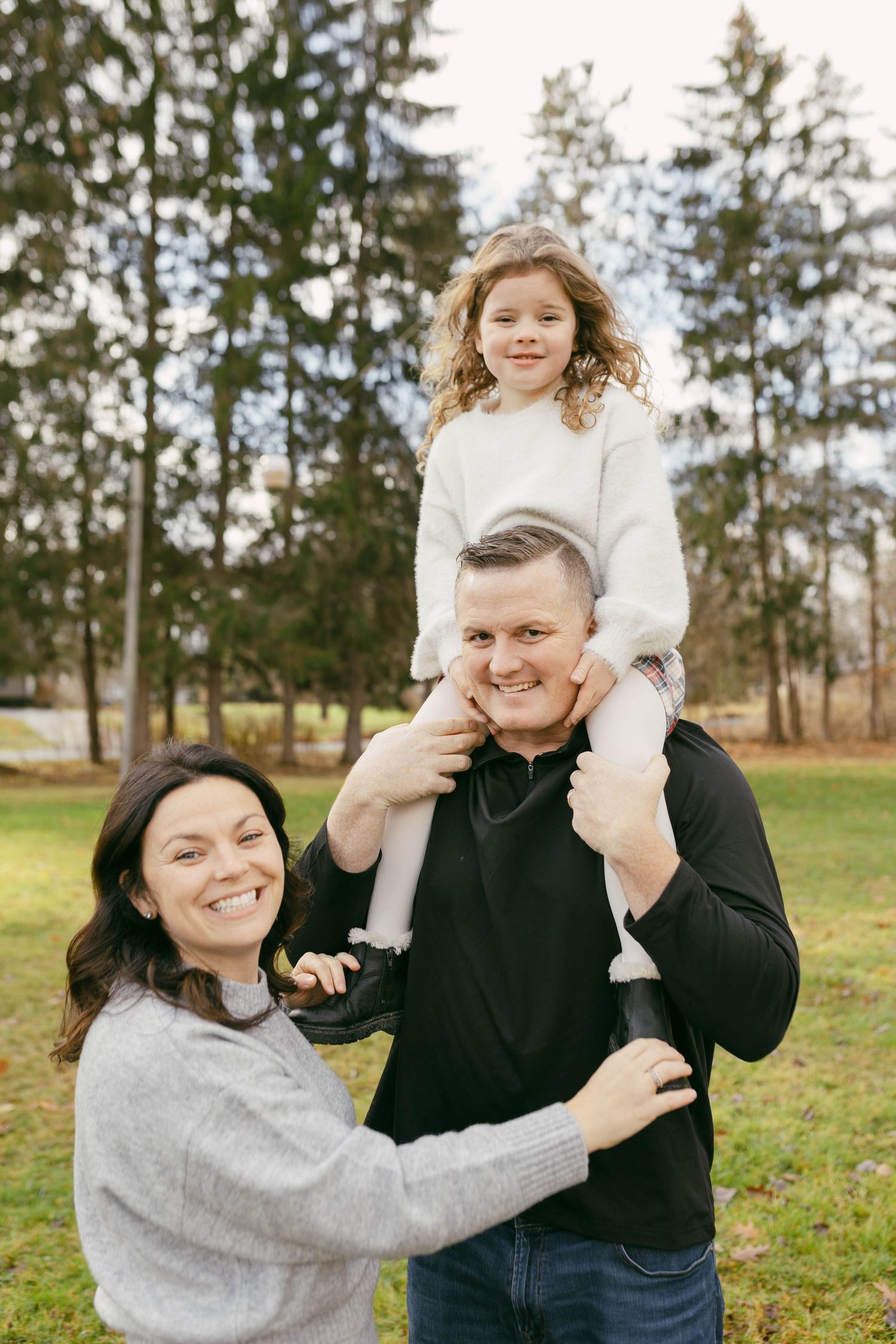 Happy family of three posing outdoors in a park, father carrying daughter on shoulders, mother smiling beside them.