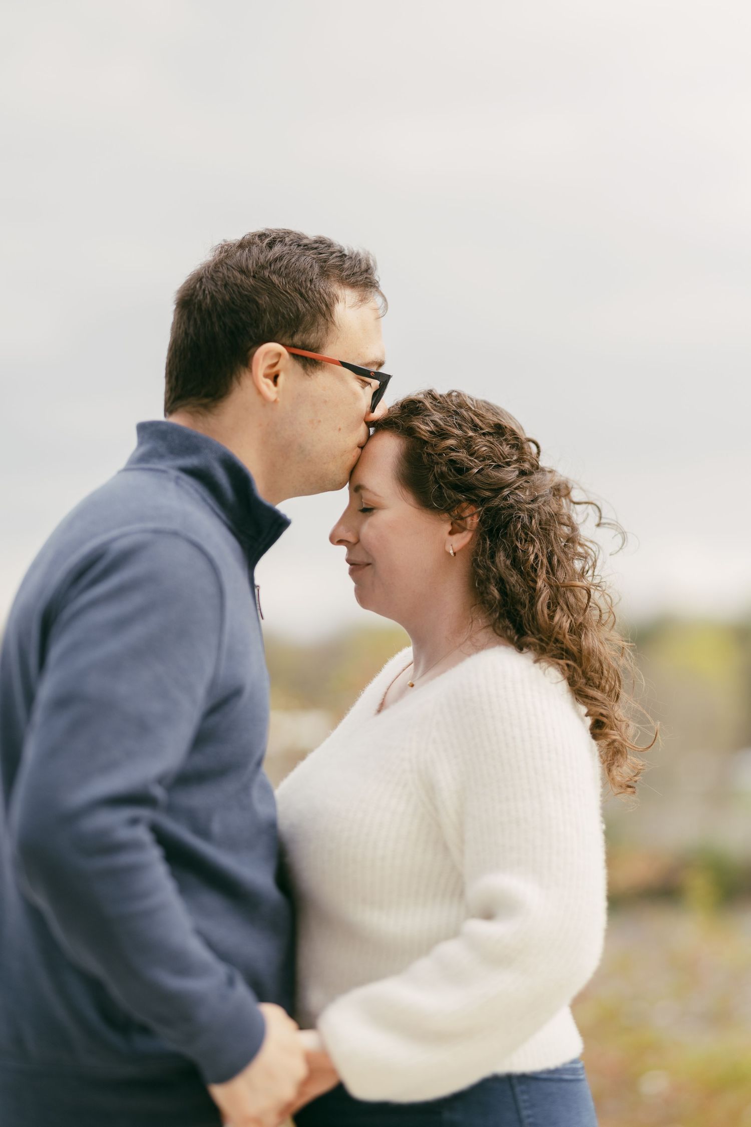 A couple shares a tender forehead kiss outdoors, man in blue jacket and woman in white sweater holding hands.