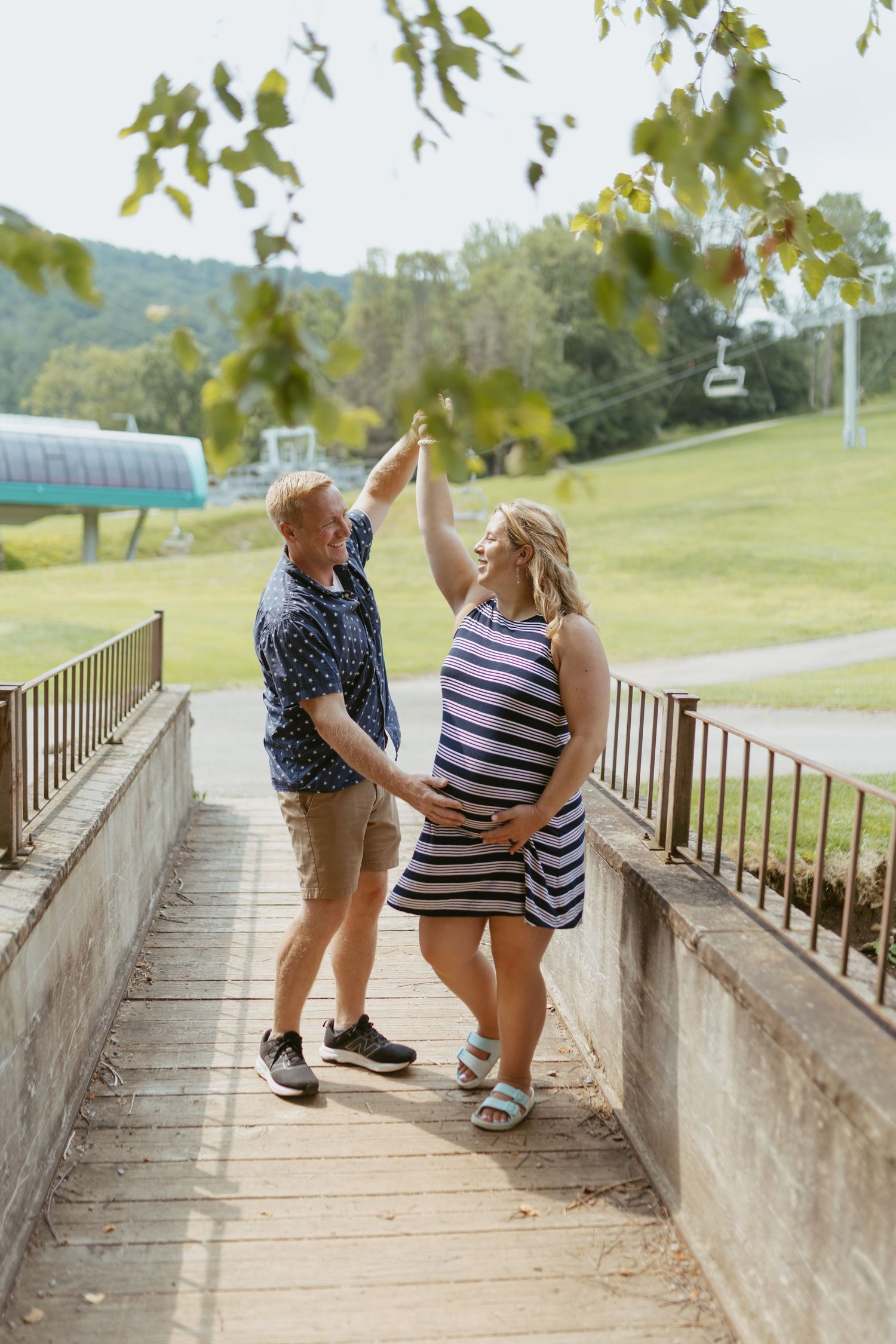 Pregnant couple dancing outdoors on a bridge, man spinning woman in striped dress, green hills and ski lift in background.
