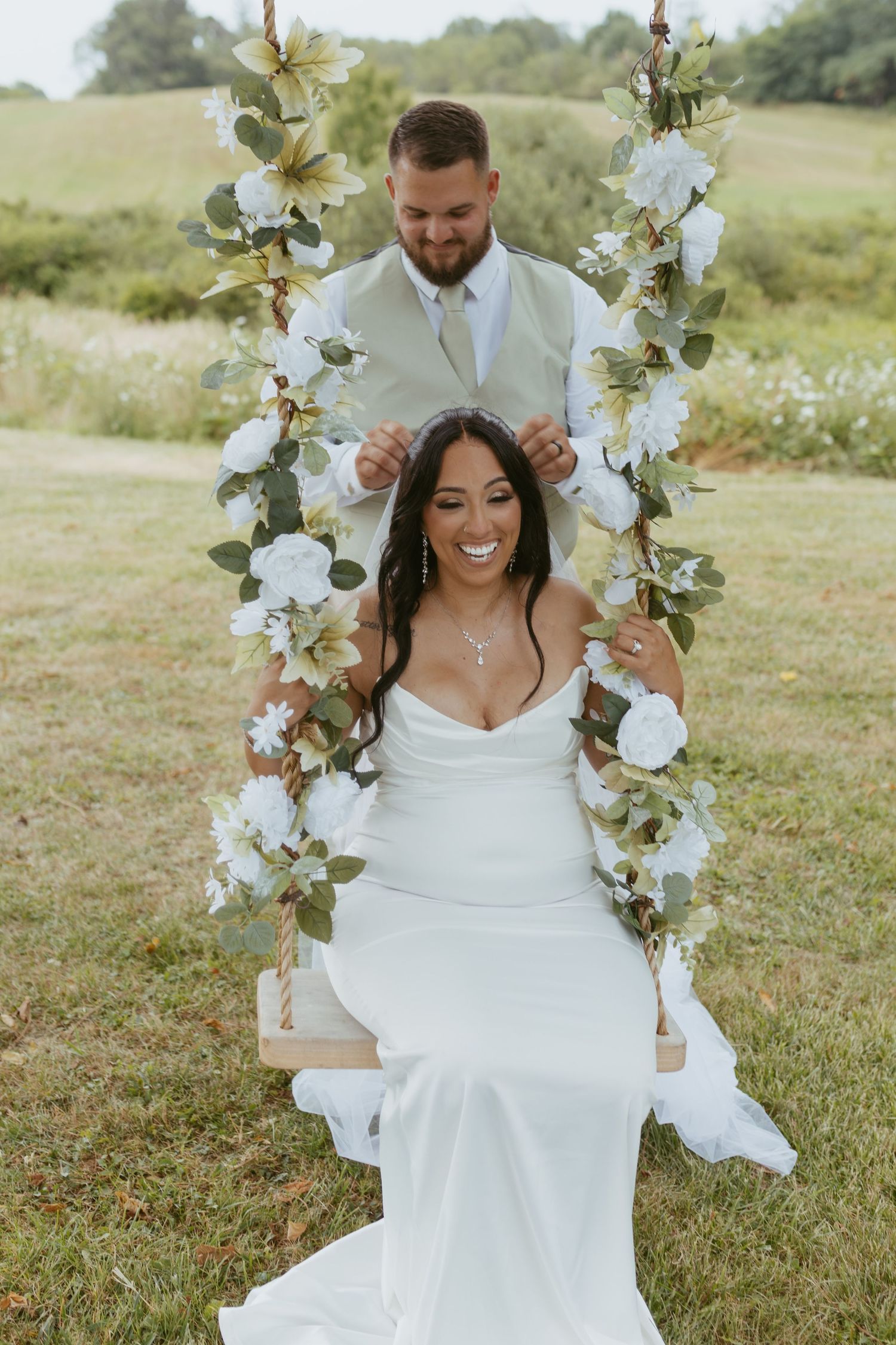 Bride in white gown sits on floral swing while groom in ivory suit stands behind her at outdoor wedding.