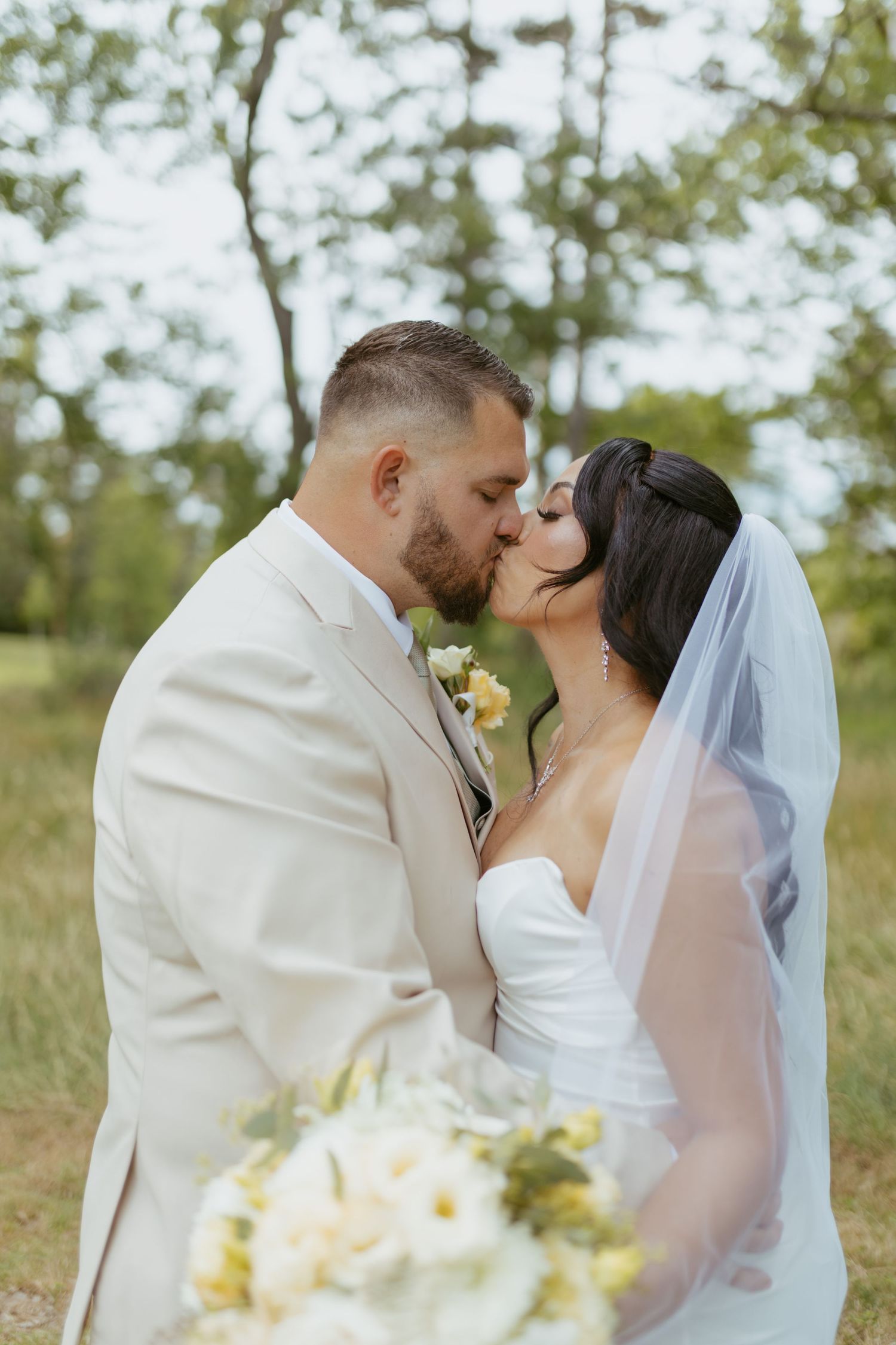 Bride and groom share a romantic kiss outdoors, bride in white gown with veil holding bouquet, groom in cream suit.