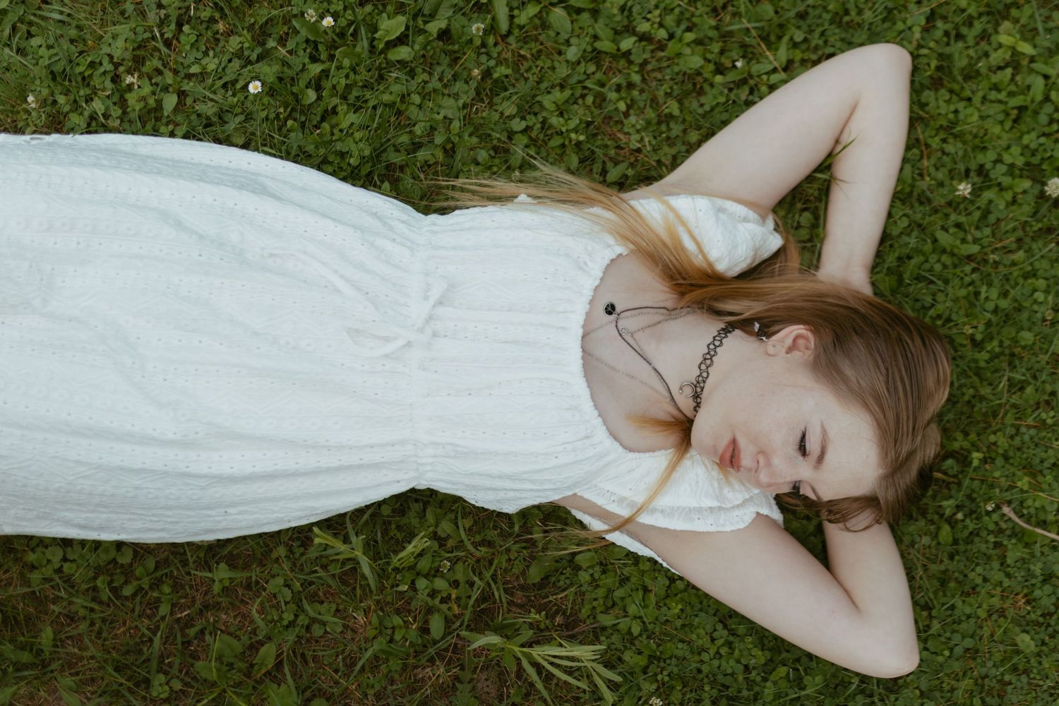Young woman in white dress lying on green grass with hands behind her head, eyes closed, relaxing outdoors.