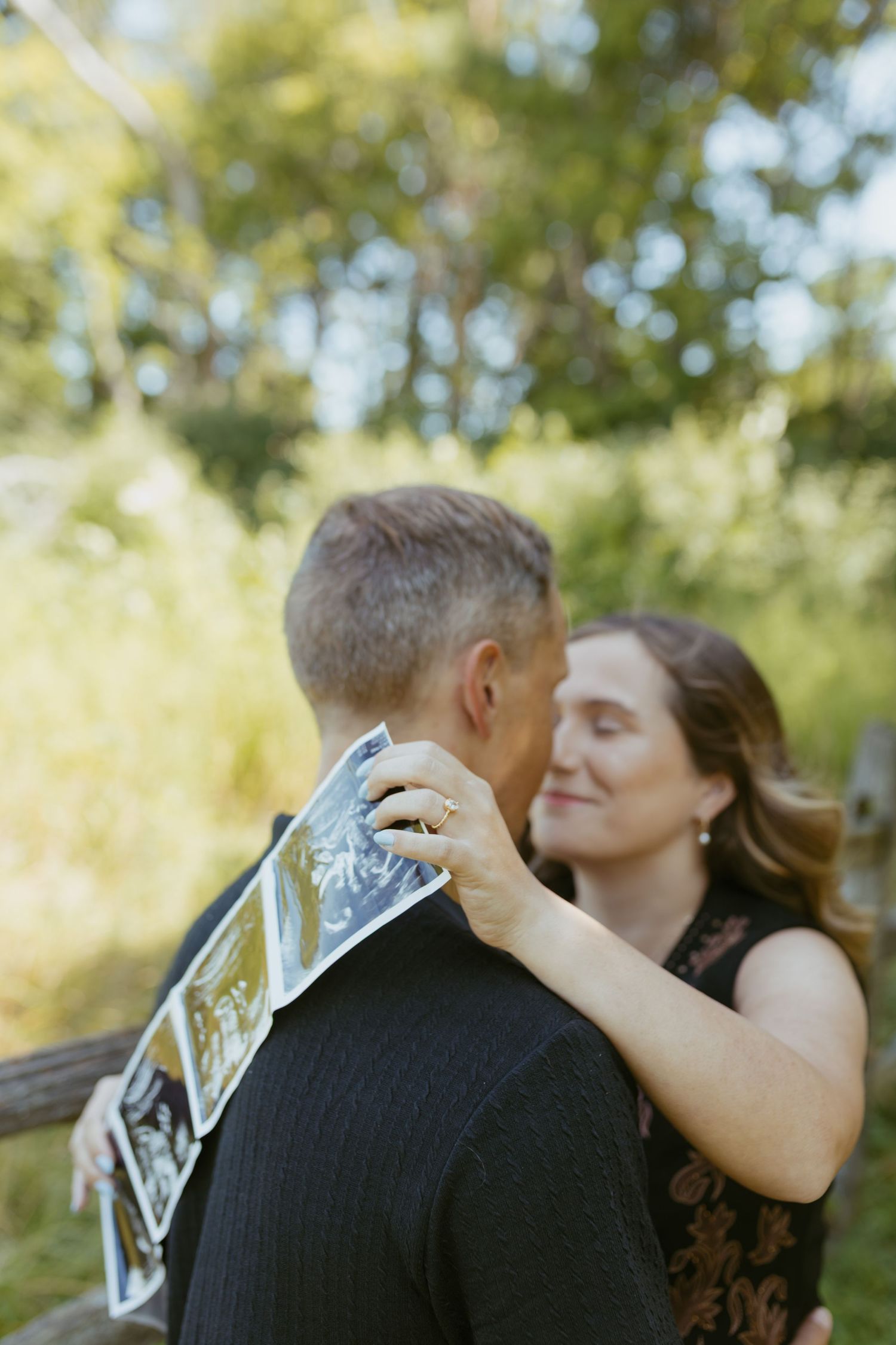 Couple embracing outdoors holding ultrasound photos, announcing pregnancy near a wooden fence with lush green trees.