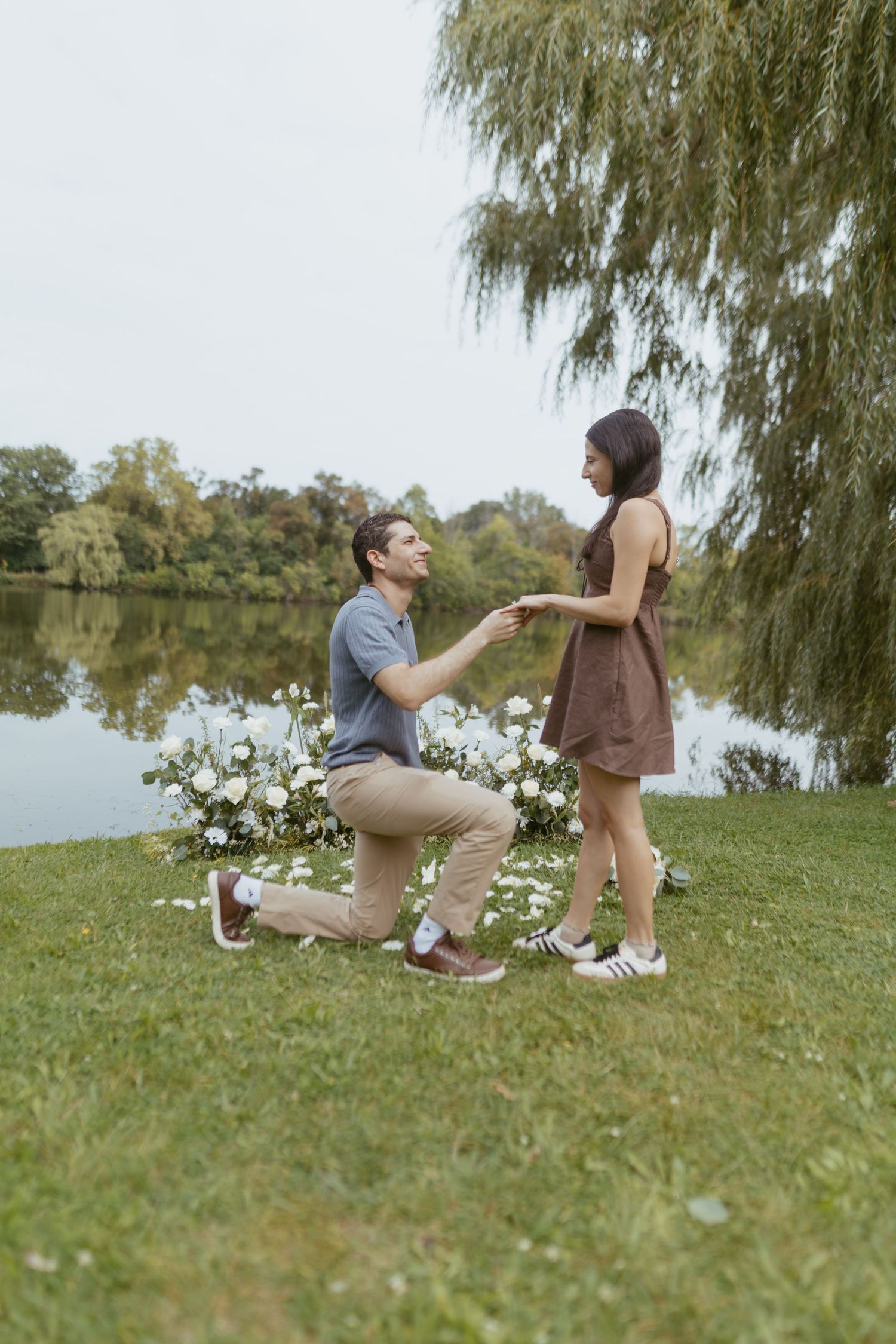 Man proposes on one knee to woman by a serene lake, surrounded by white flowers and weeping willow trees.