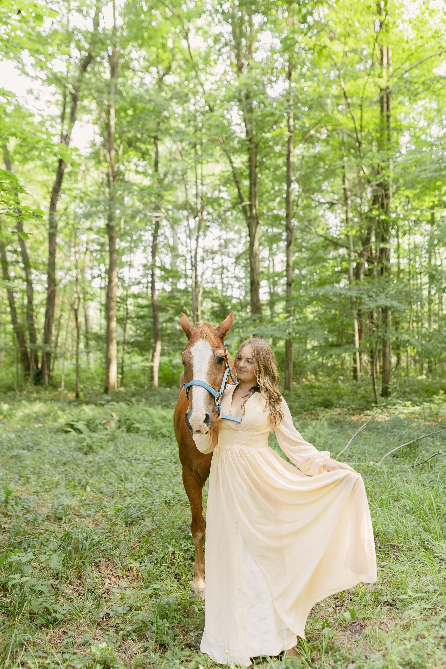 Someone in a cream-colored dress poses with a horse in a green forest setting.